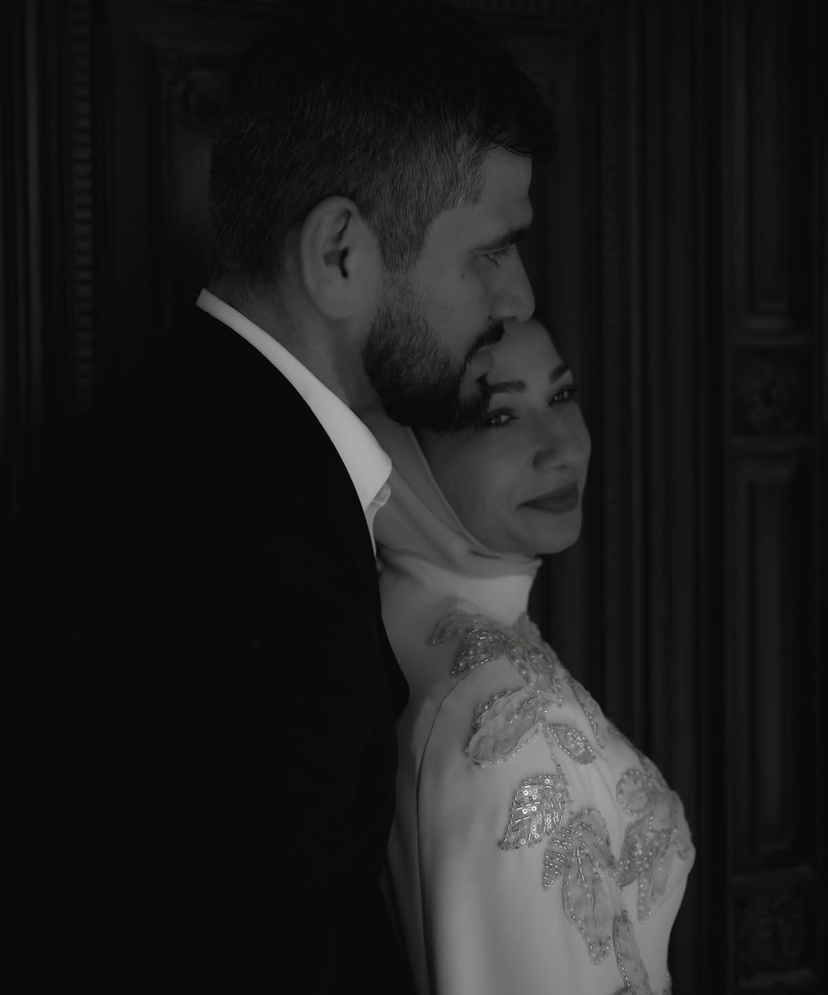 Black and white photo of a couple in formal attire, woman in embroidered dress leaning against man in suit.
