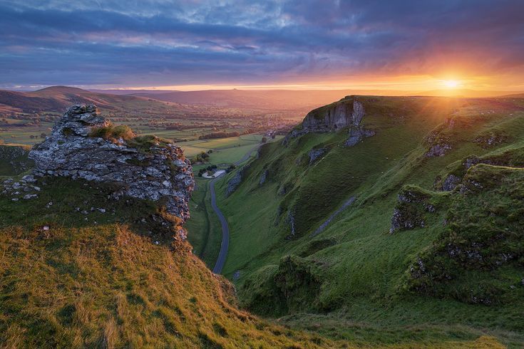 Stunning sunset over a lush green valley with dramatic rocky cliffs and a winding road in the Peak District.