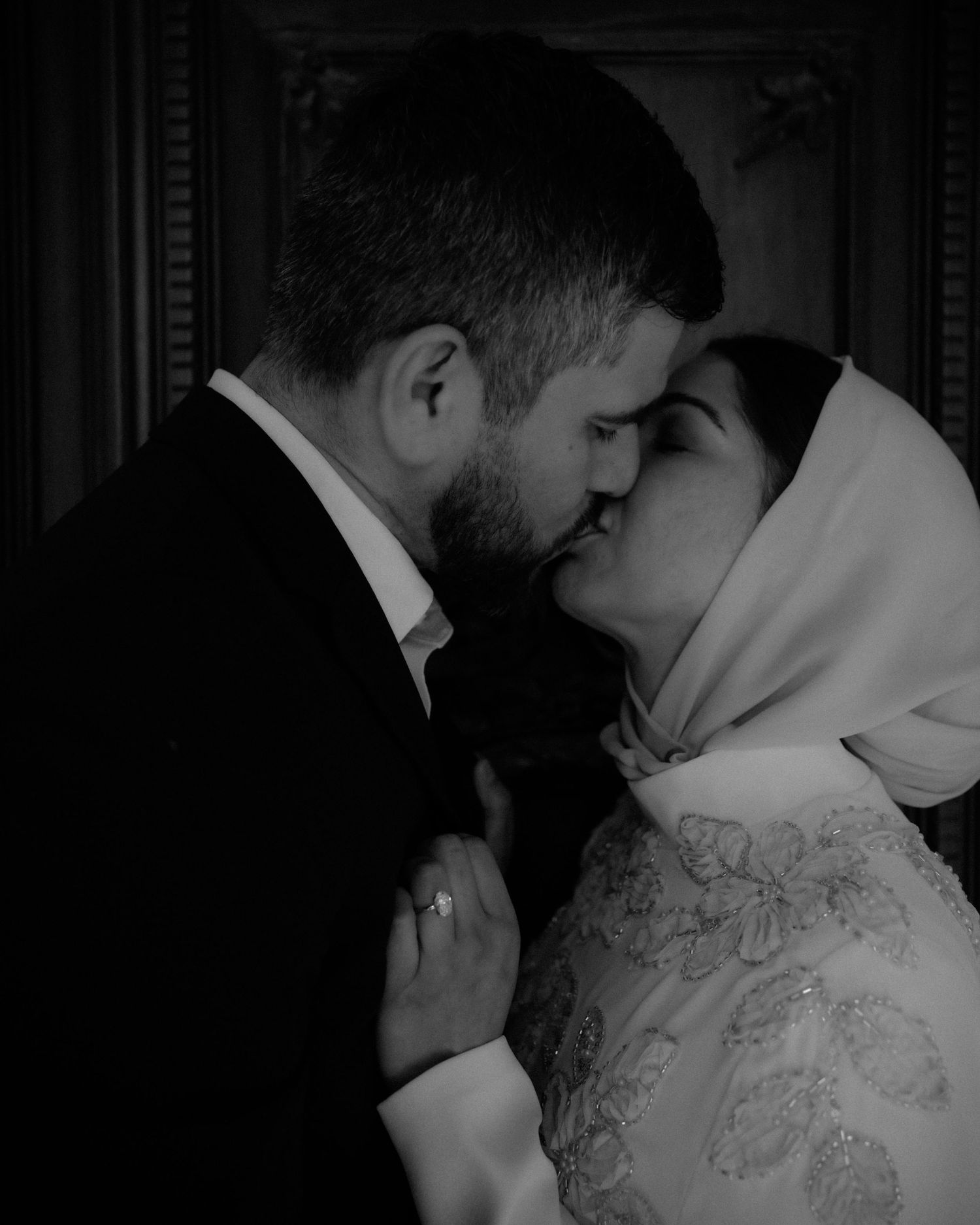 Black and white photo of a couple sharing a romantic kiss, bride in hijab and gown, groom in suit.