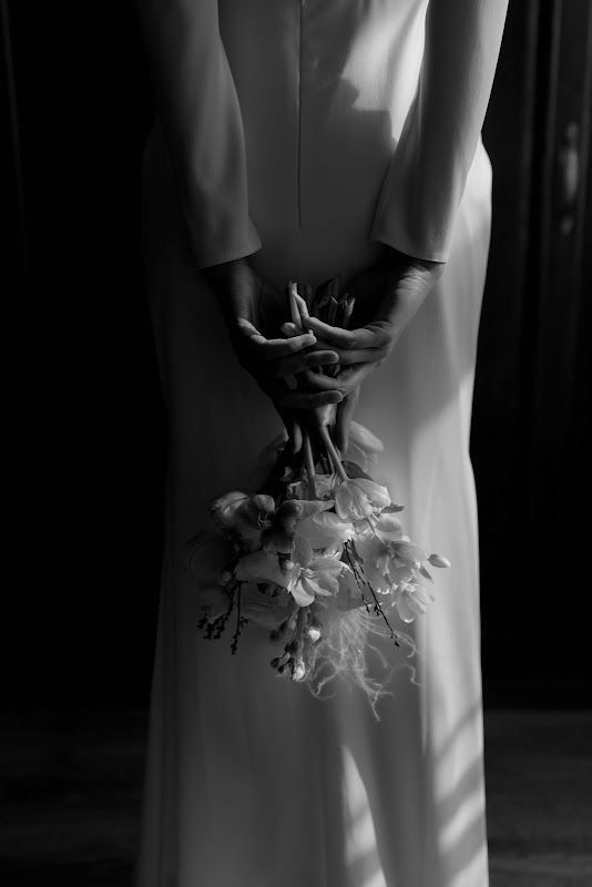 Bride in white gown holds small floral bouquet behind her back, black and white wedding photo.