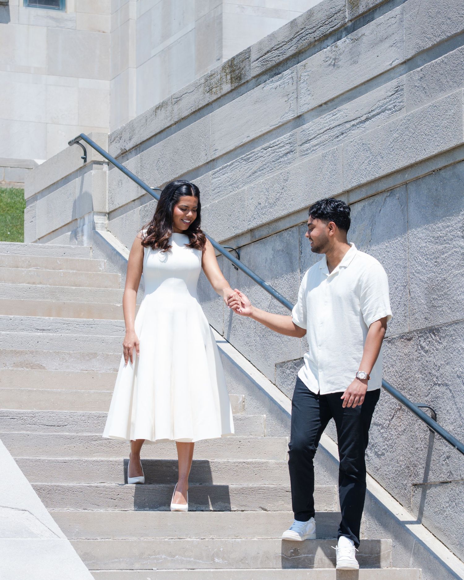 Couple in white outfits holding hands on stone steps of a grand building on a sunny day.