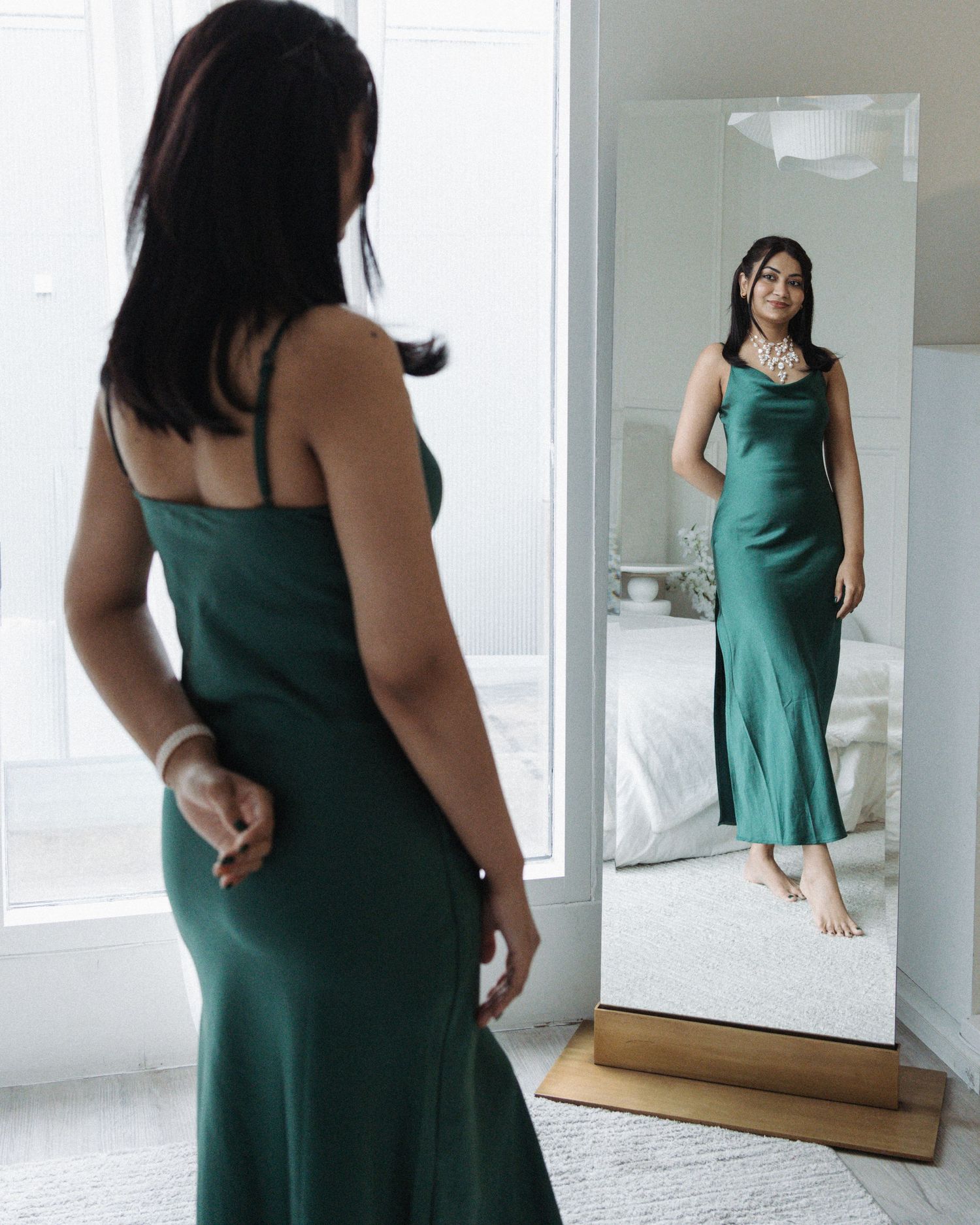 Woman in elegant green satin slip dress posing before a full-length mirror in a bright bedroom.