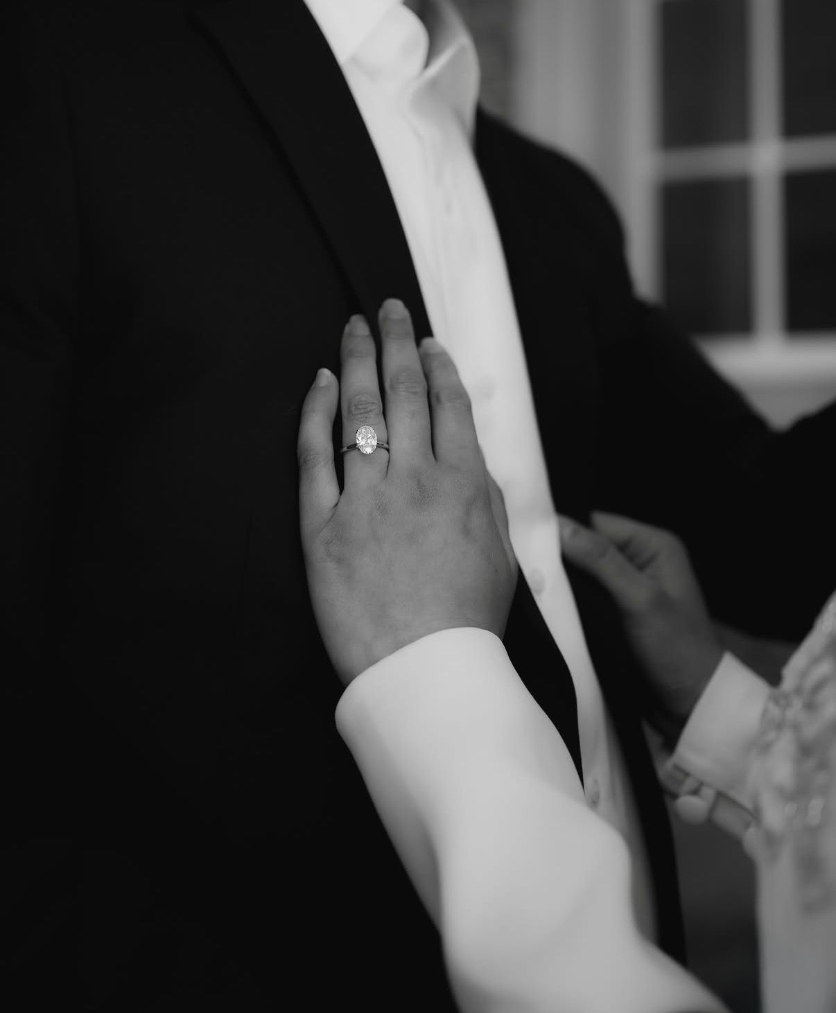 Black and white close-up of a bride's hand with elegant engagement ring resting on groom's suit lapel.