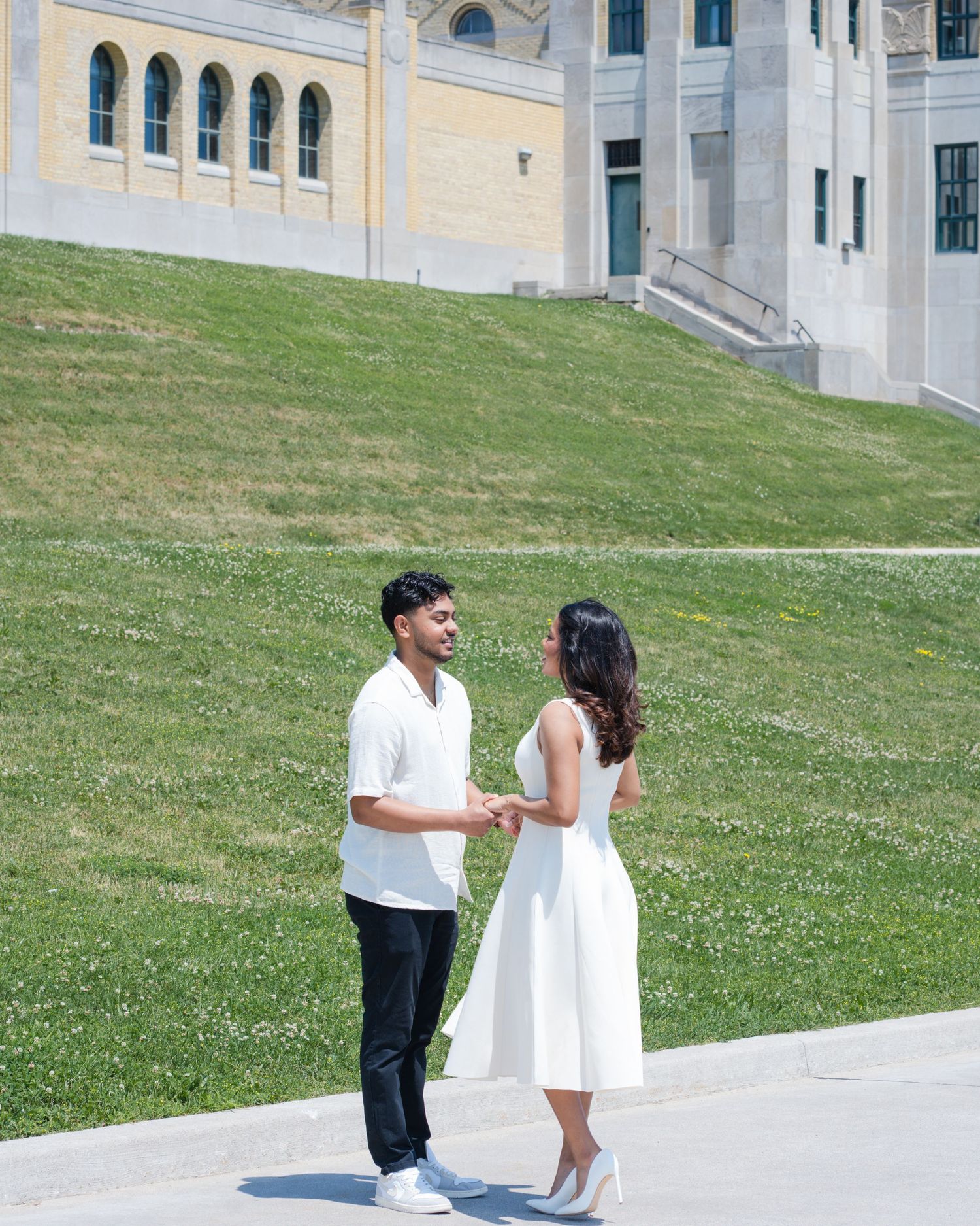 Couple holding hands in white outfits on a sunny day outside a stone building with green lawn.
