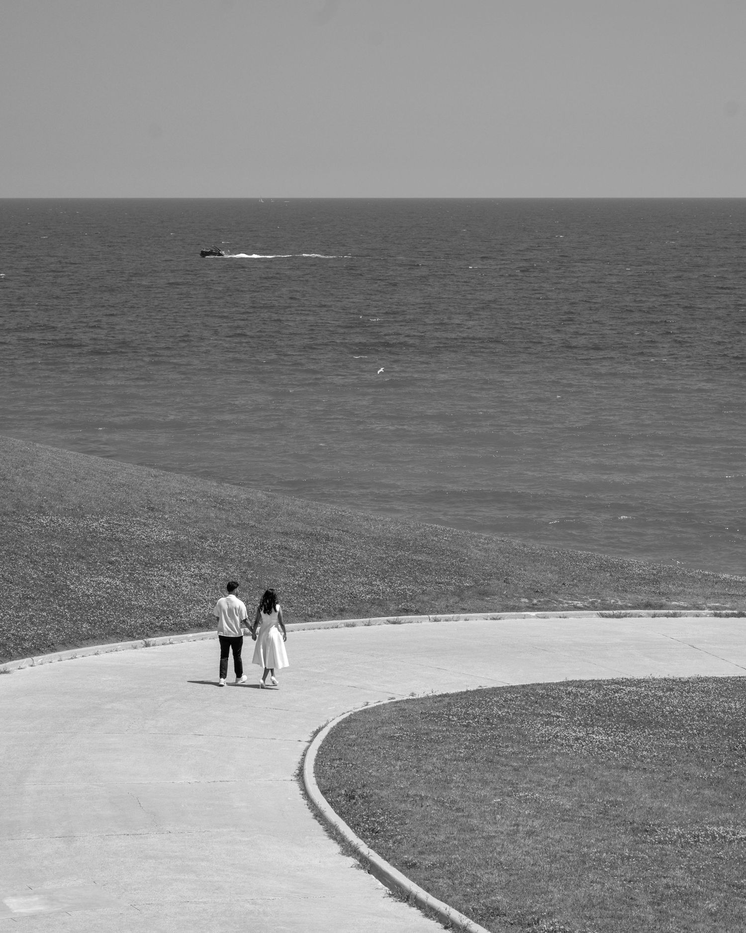 Couple walking along a curved seaside promenade with ocean horizon in black and white photography.