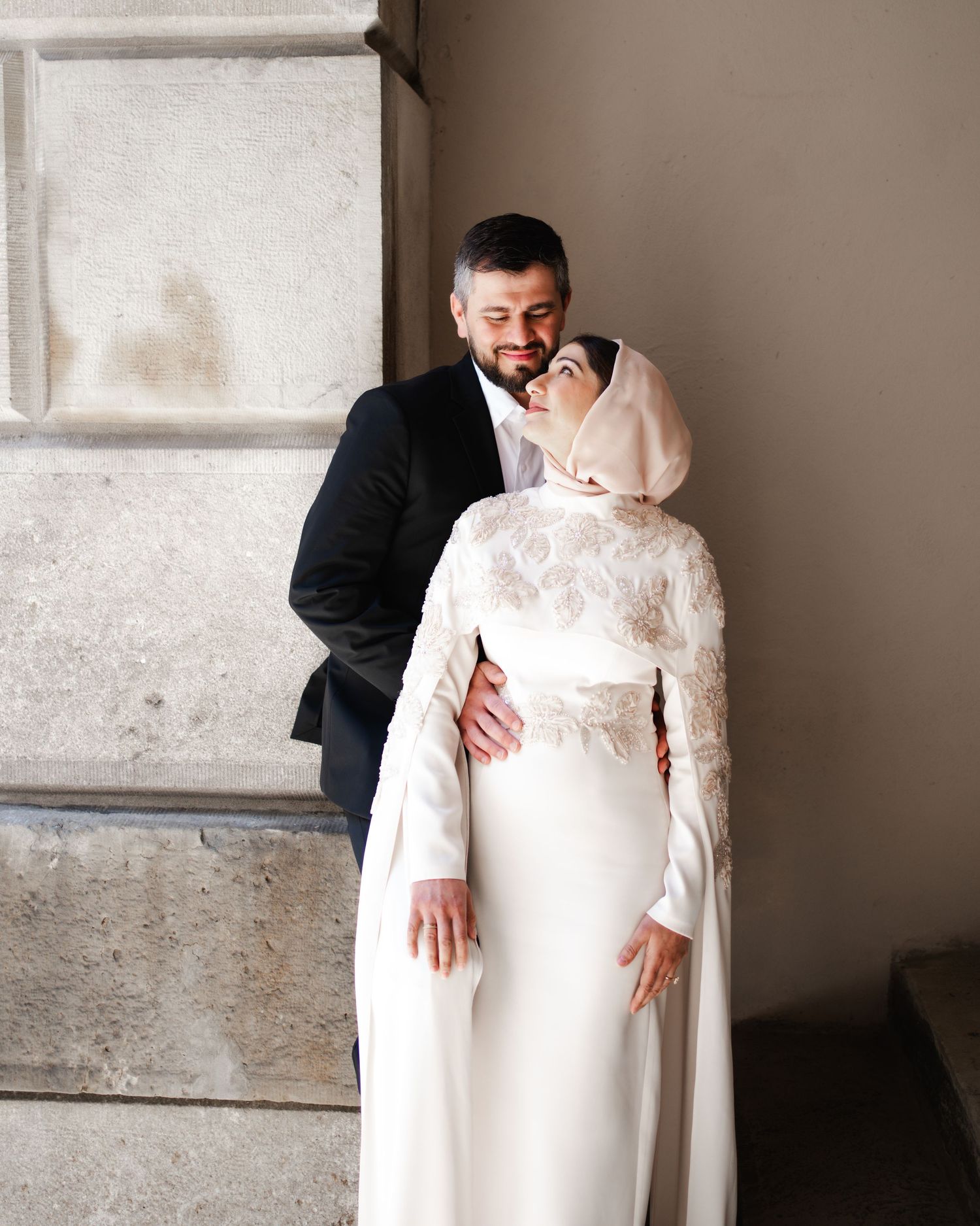 A groom in a black suit kisses his bride wearing a white hijab and elegant lace wedding gown.