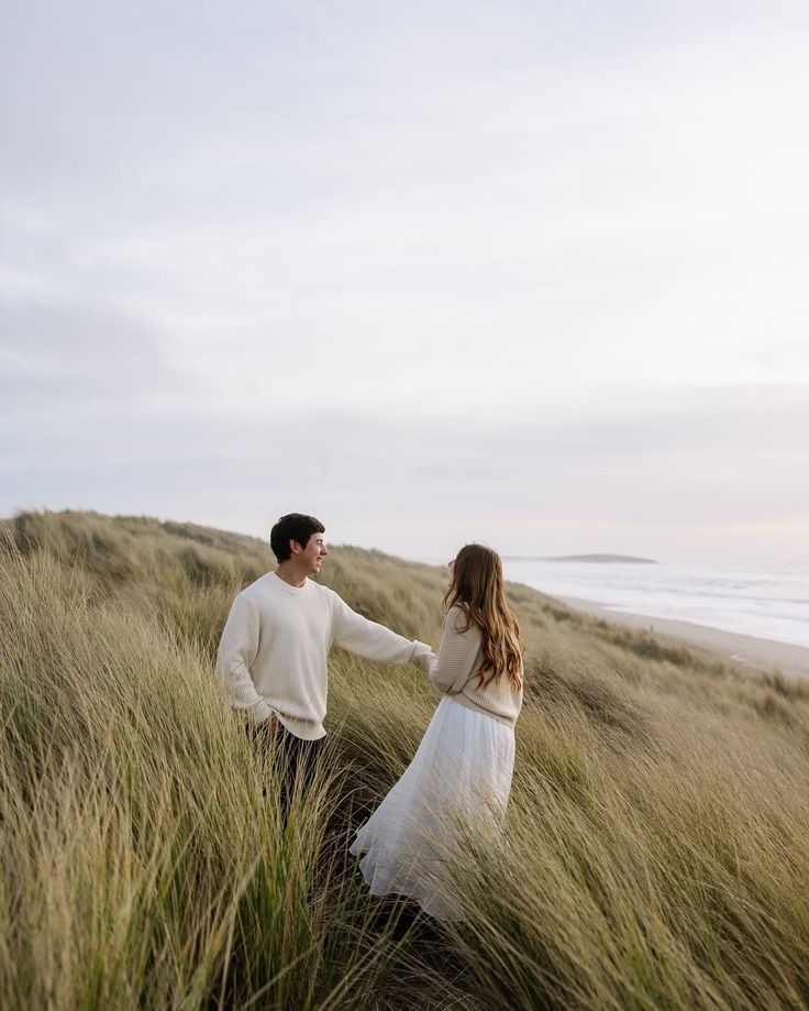Couple walking through tall windswept grass on coastal sand dunes at sunset with ocean view in background.