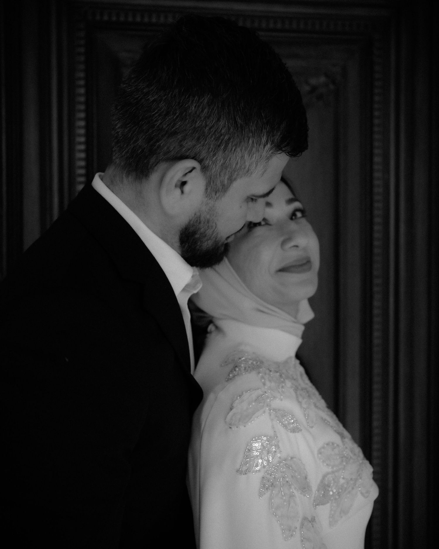 Black and white photo of a couple in wedding attire, woman in hijab and embroidered gown, man in suit embracing her.