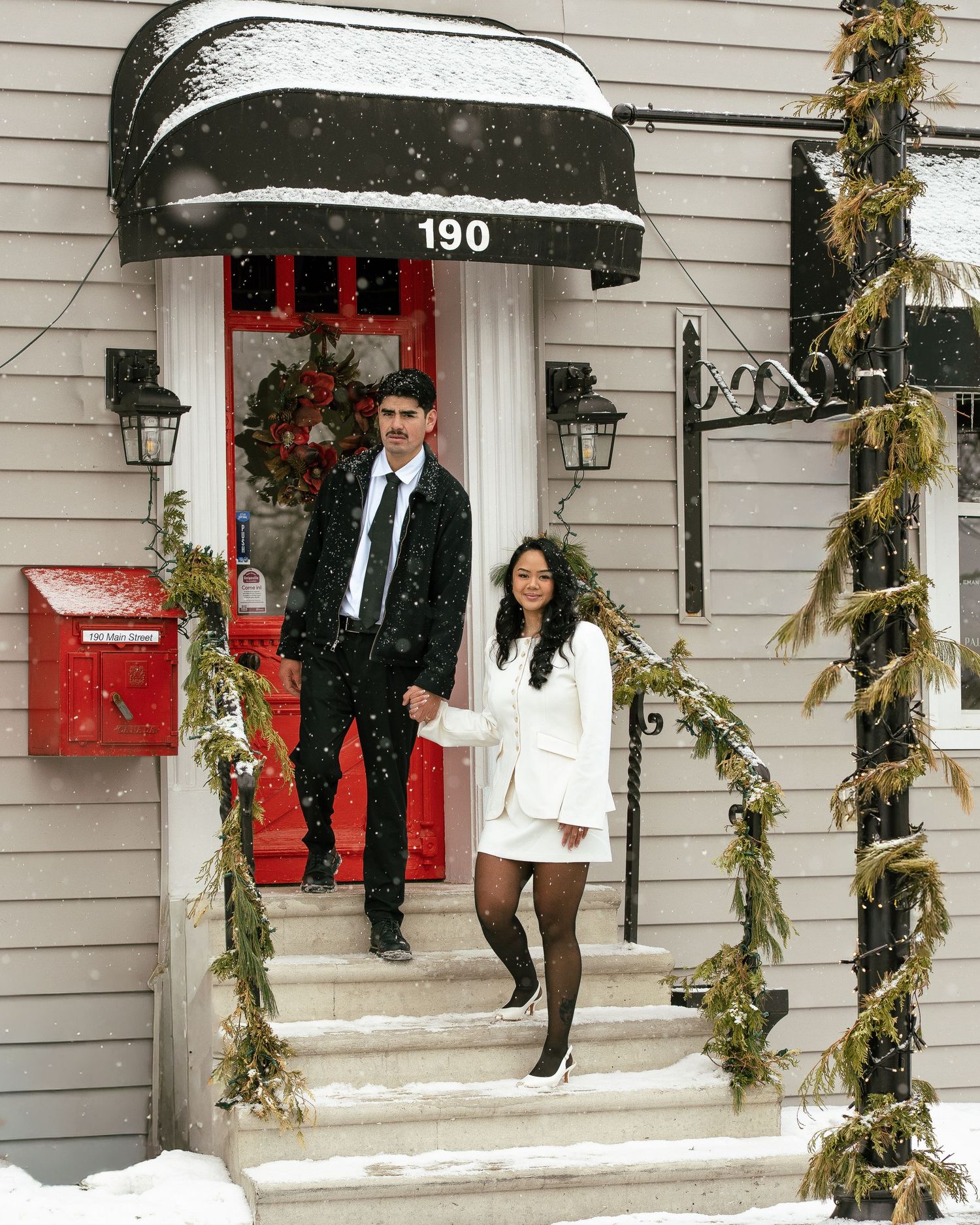 Couple in formal attire holding hands on snowy steps at address 190, decorated with garland and a red door.