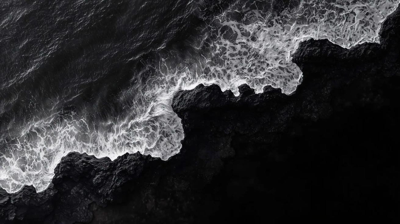 Aerial black and white view of ocean waves crashing against dark rocky coastline with white sea foam.