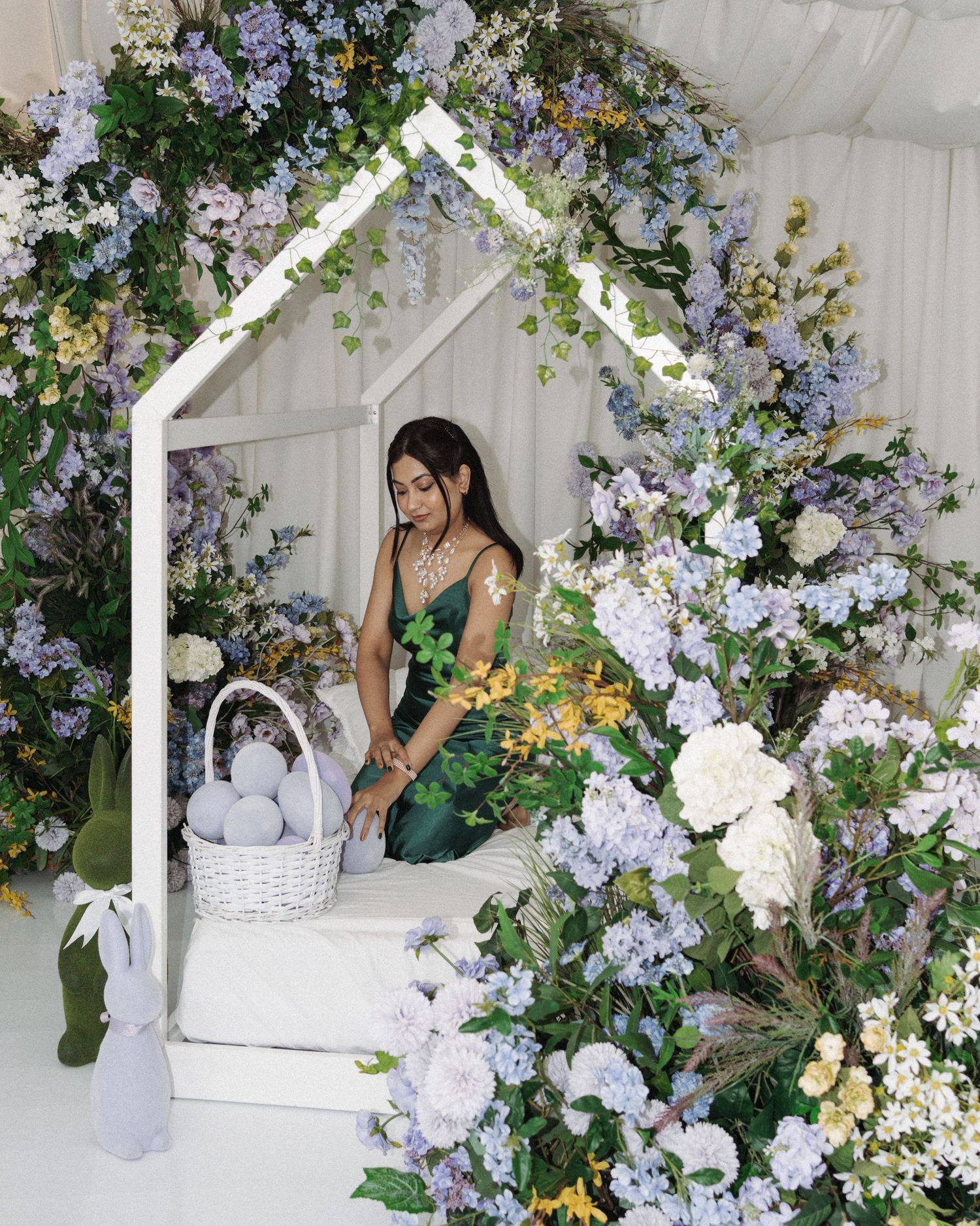 Woman in green dress sitting in white house frame surrounded by purple and white floral arrangement with Easter basket.