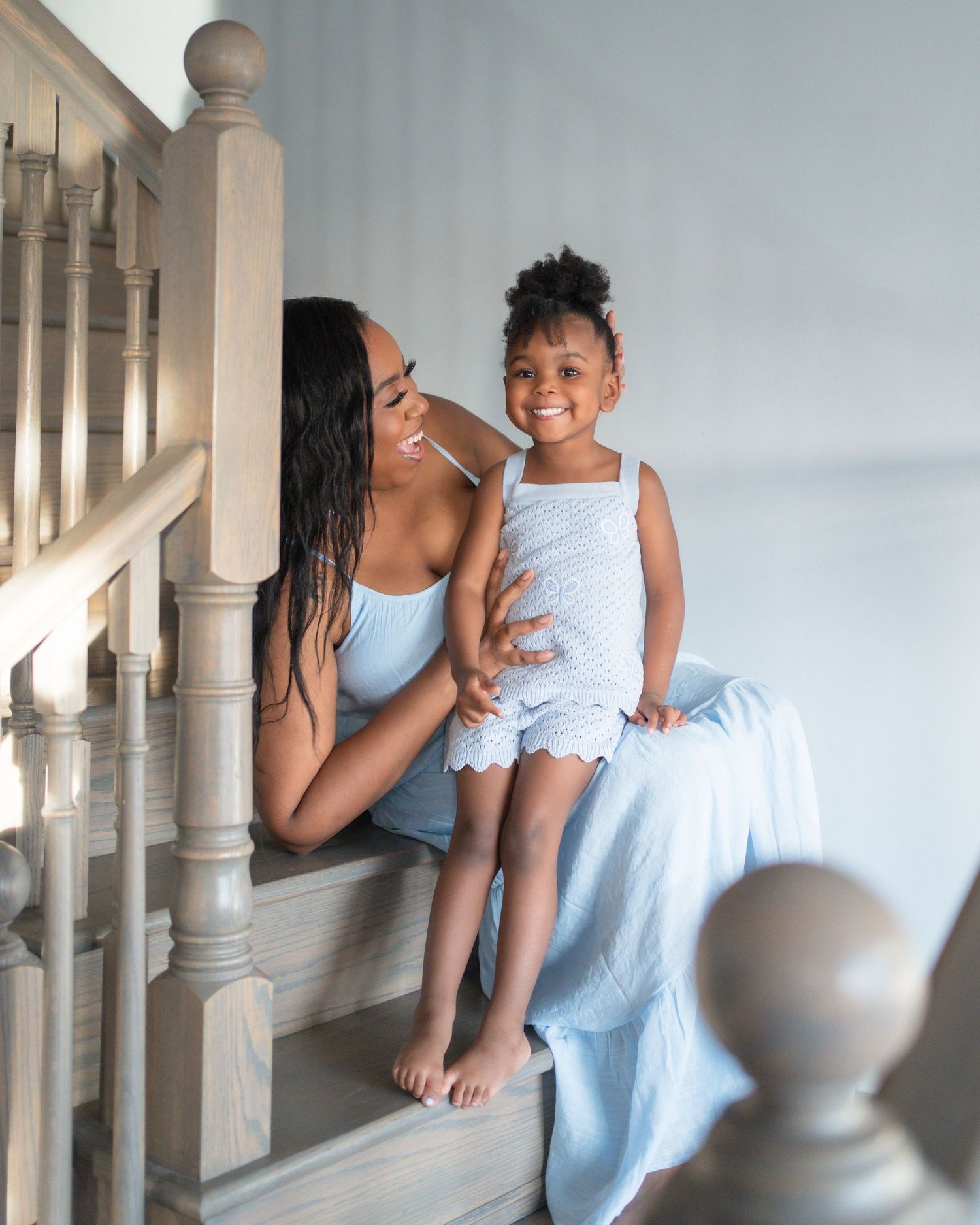 A mother and young daughter sit together on wooden stairs, both wearing matching light blue outfits, smiling warmly.