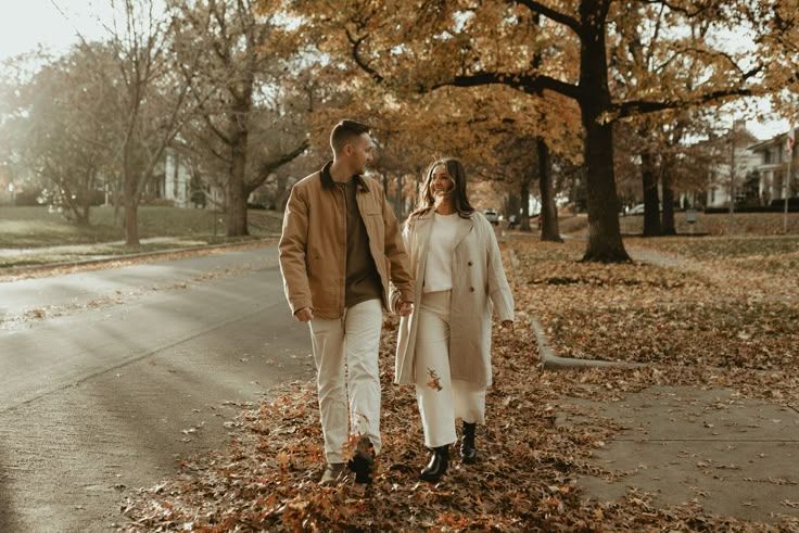 Couple walking hand-in-hand through a sunlit autumn park, wearing cozy beige coats surrounded by golden fallen leaves.