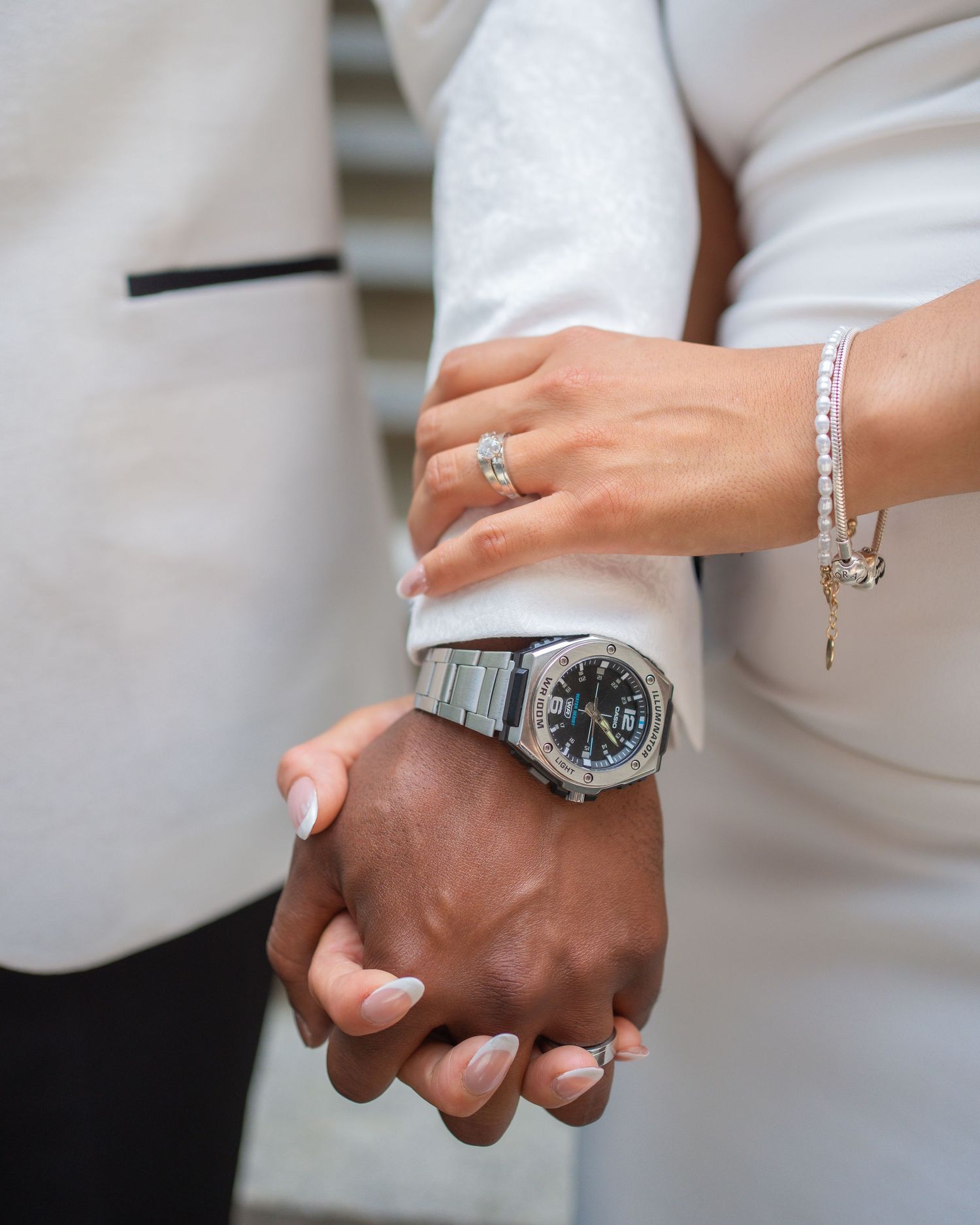 Close-up of interracial couple holding hands, woman wearing diamond ring and bracelet, man wearing silver luxury watch.