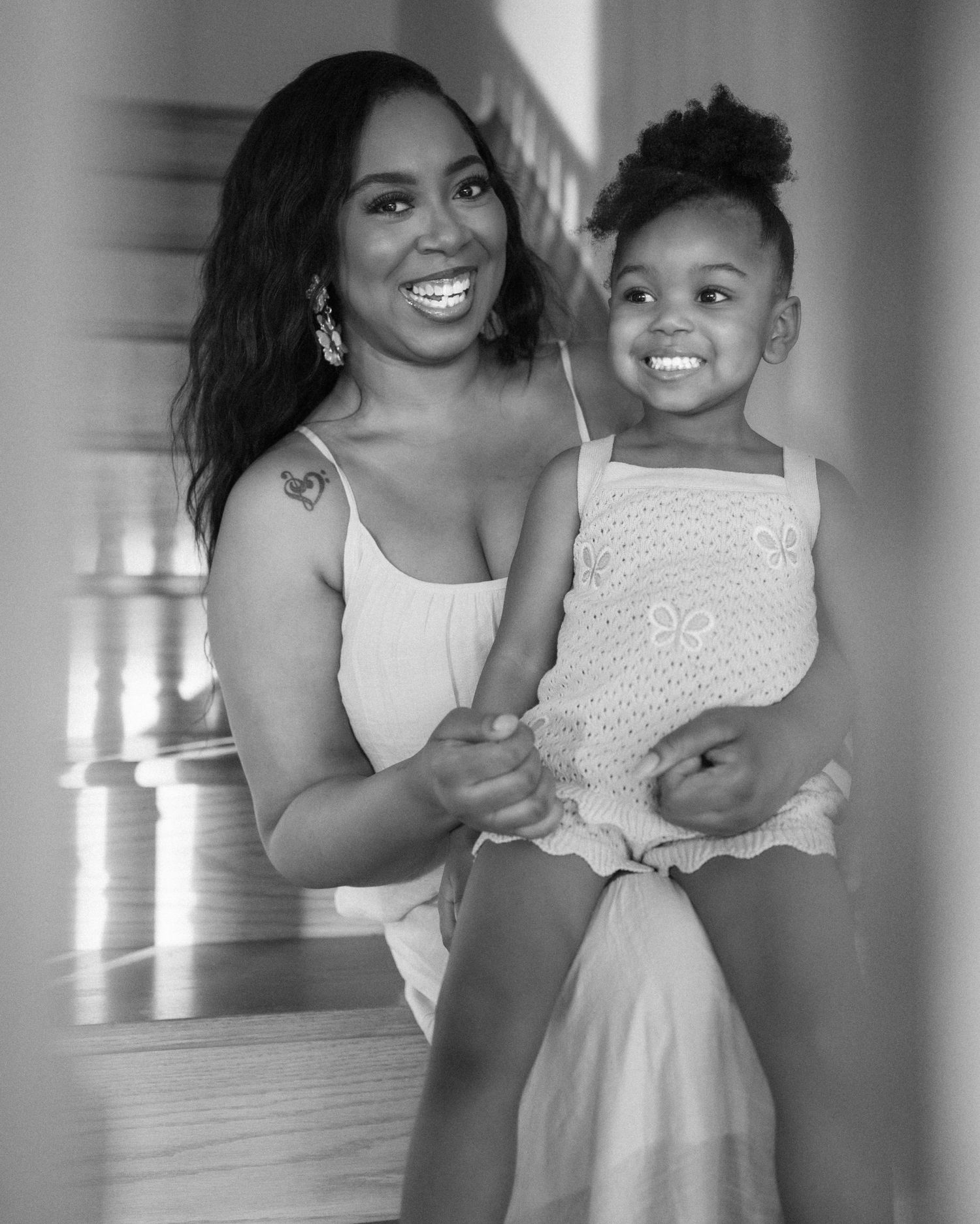 Black and white photo of a smiling woman holding a young girl, both dressed in white, posing on a staircase.
