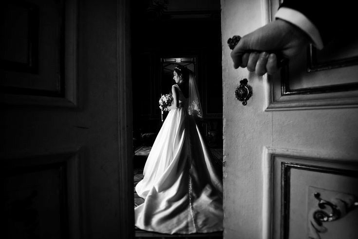 Black and white photo of a bride in a white gown holding bouquet, framed by a doorway with a hand on the knob.