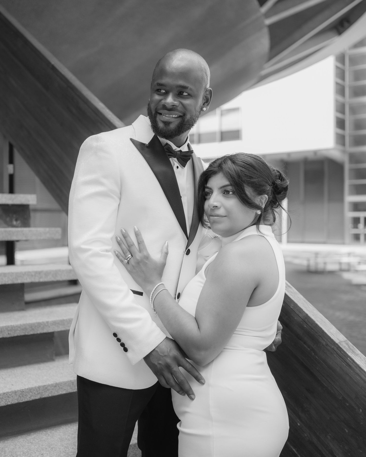 Black and white photo of a smiling couple in wedding attire embracing outdoors near modern architecture.