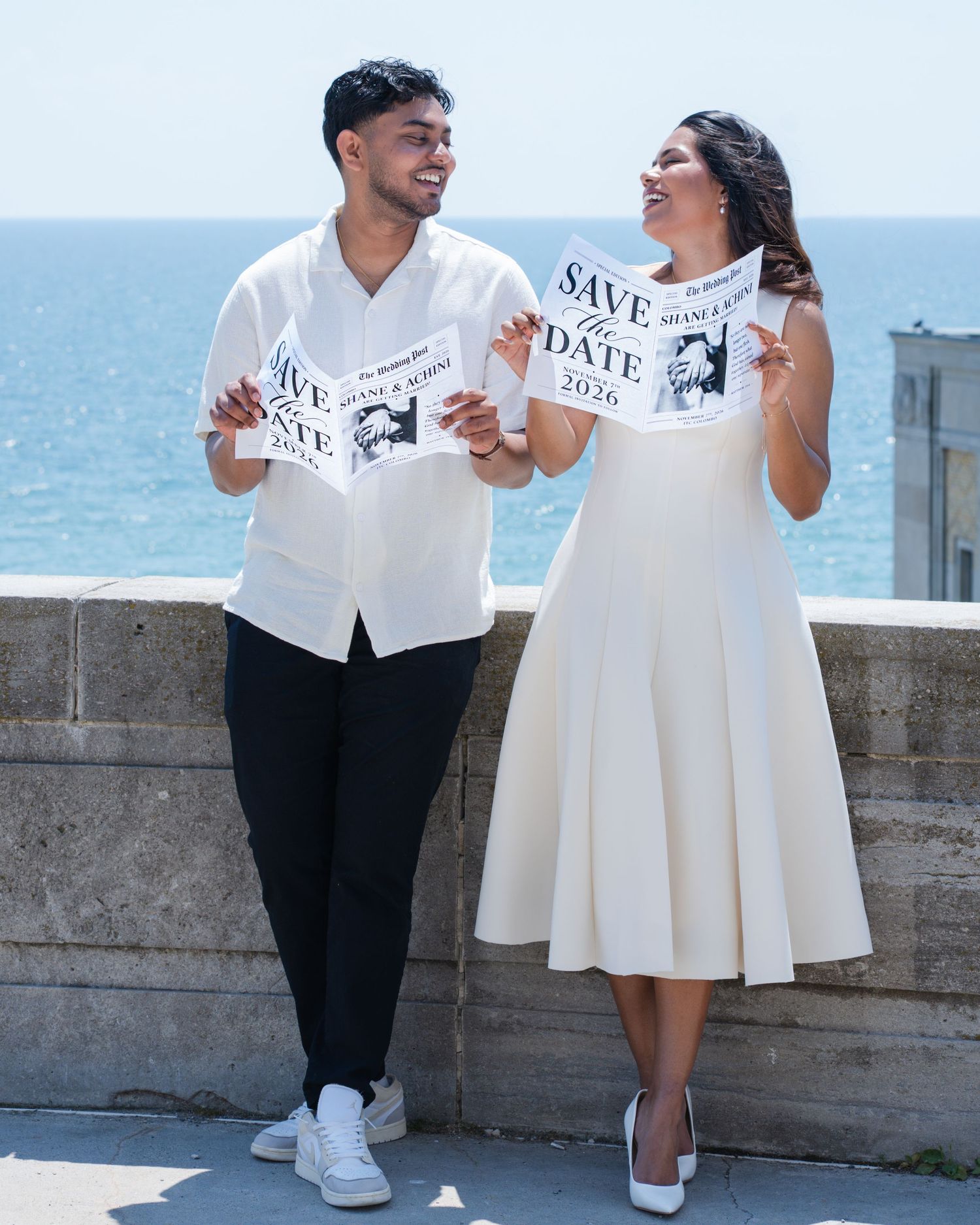 Couple holding Save the Date cards by the ocean, dressed in white outfits, celebrating their engagement.