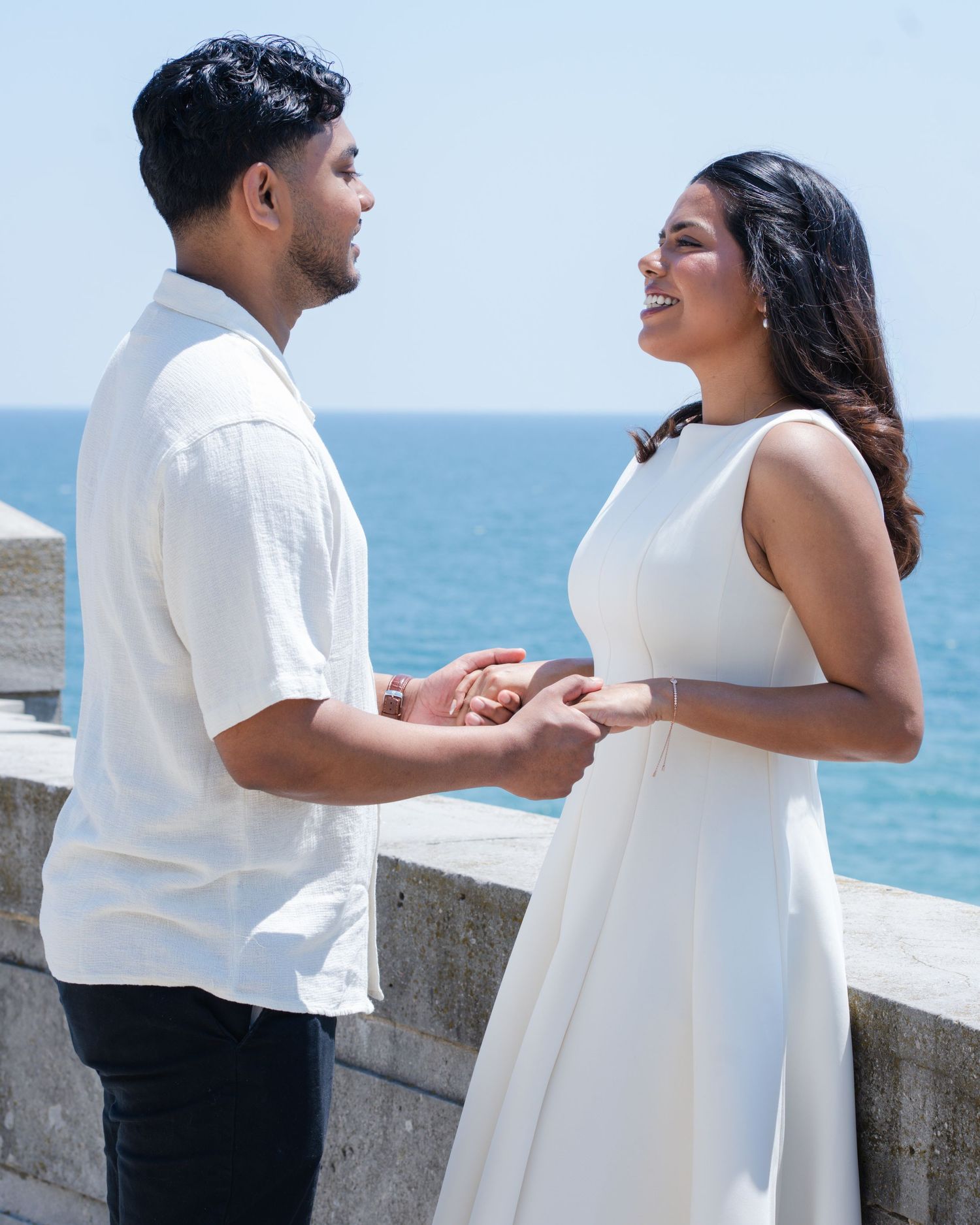 Couple in white outfits holding hands by ocean wall on sunny day.