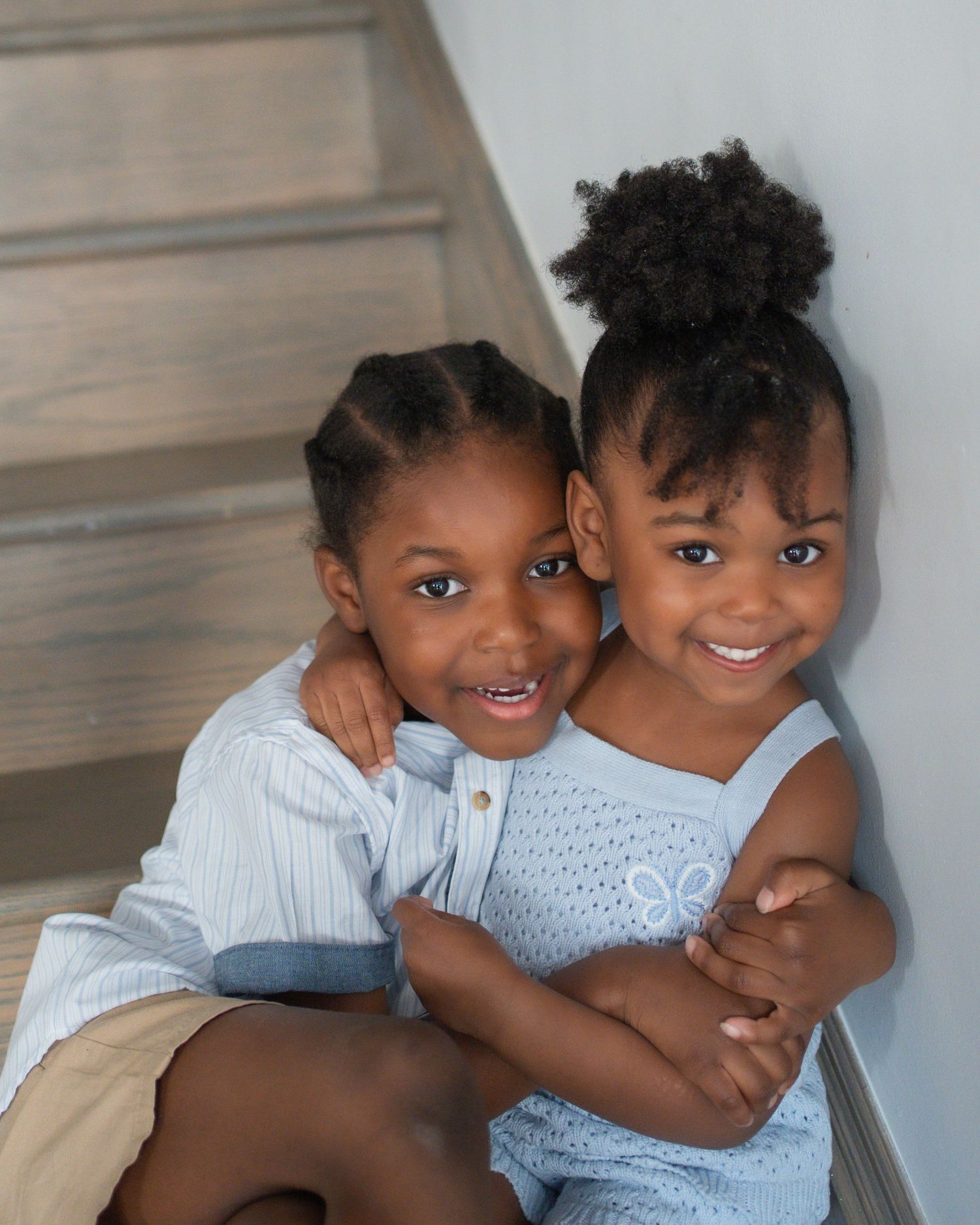 Two young Black girls hugging and smiling on stairs, wearing matching light blue outfits in a bright indoor setting.