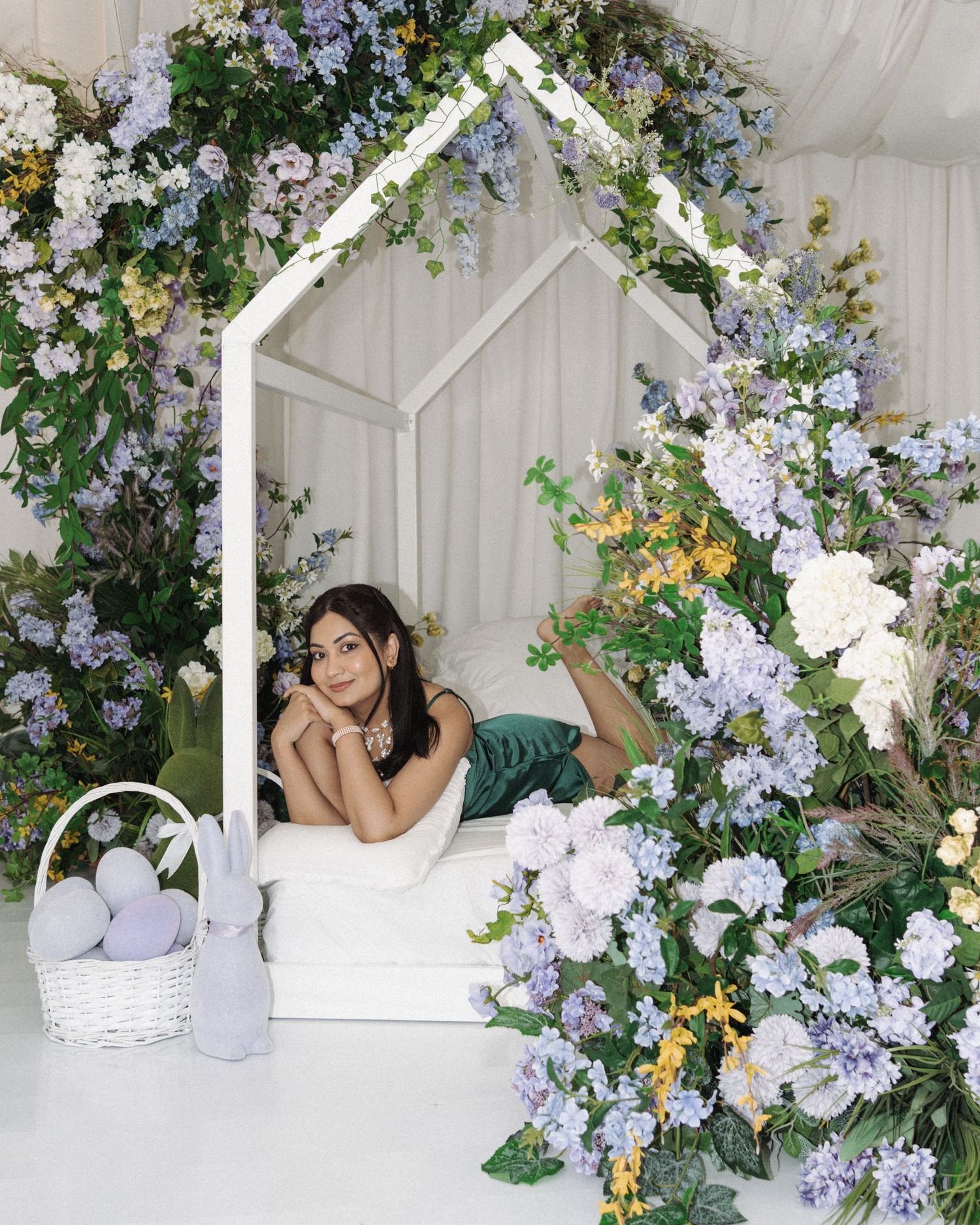 Woman posing in a floral arch bed frame surrounded by purple hydrangeas and greenery with Easter eggs nearby.