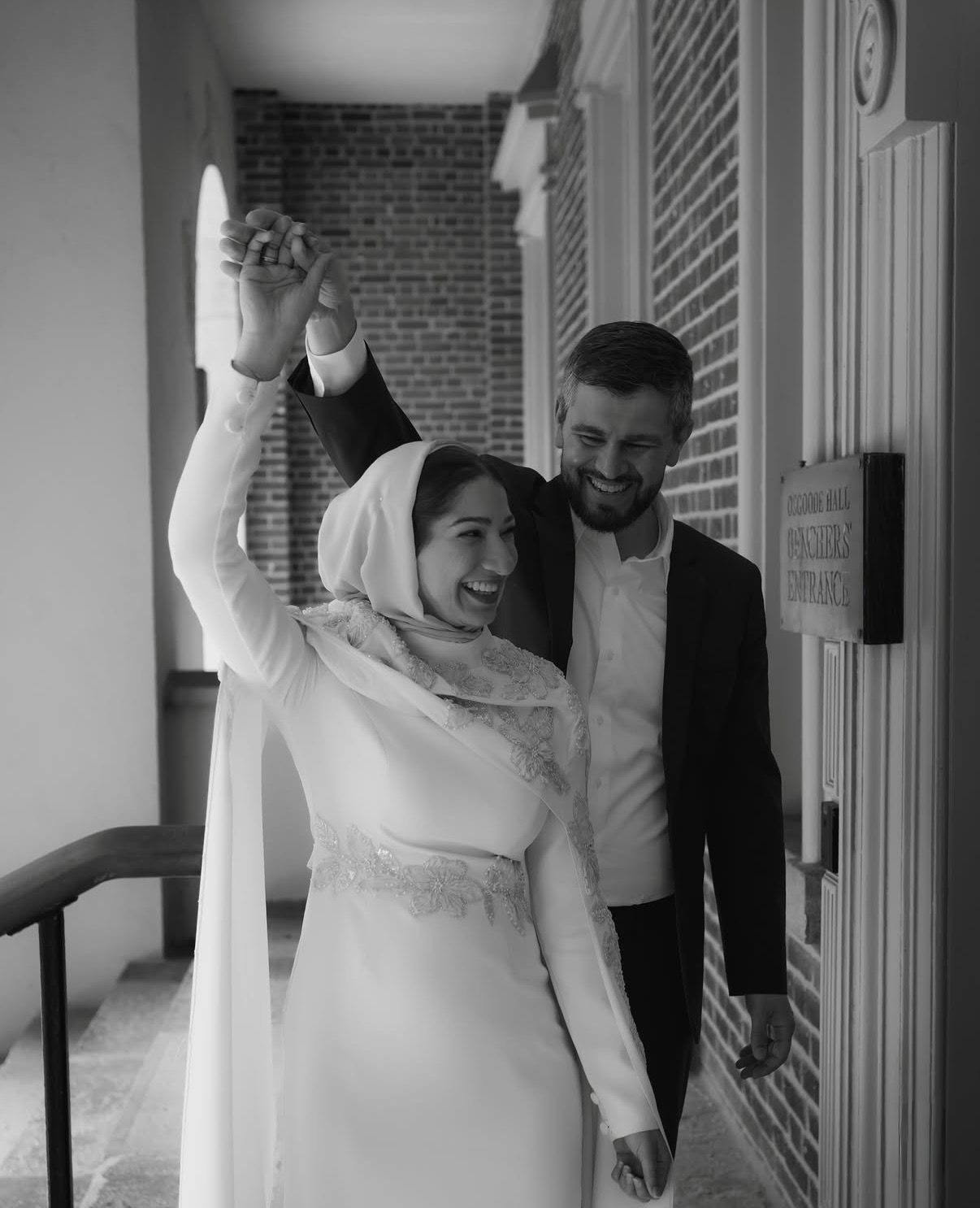 Joyful newlywed couple celebrating outside brick building, bride in hijab and white gown, groom in black suit.