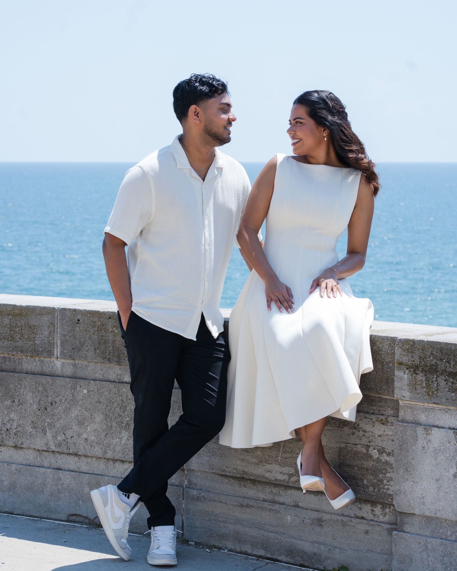 A couple in matching white outfits poses by a stone wall overlooking the ocean on a sunny day.