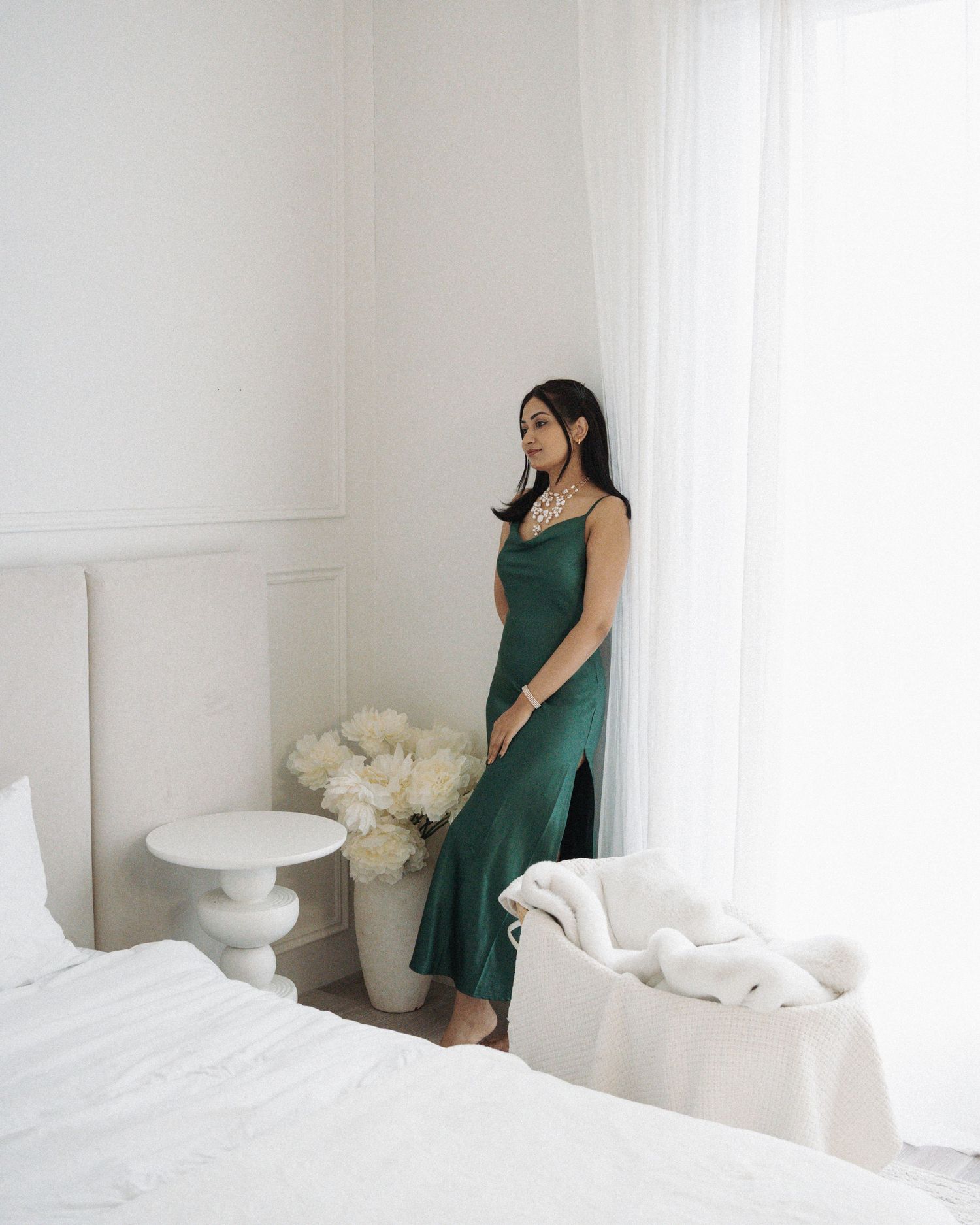 Woman in elegant green satin slip dress standing by bright white curtains in a minimalist bedroom.