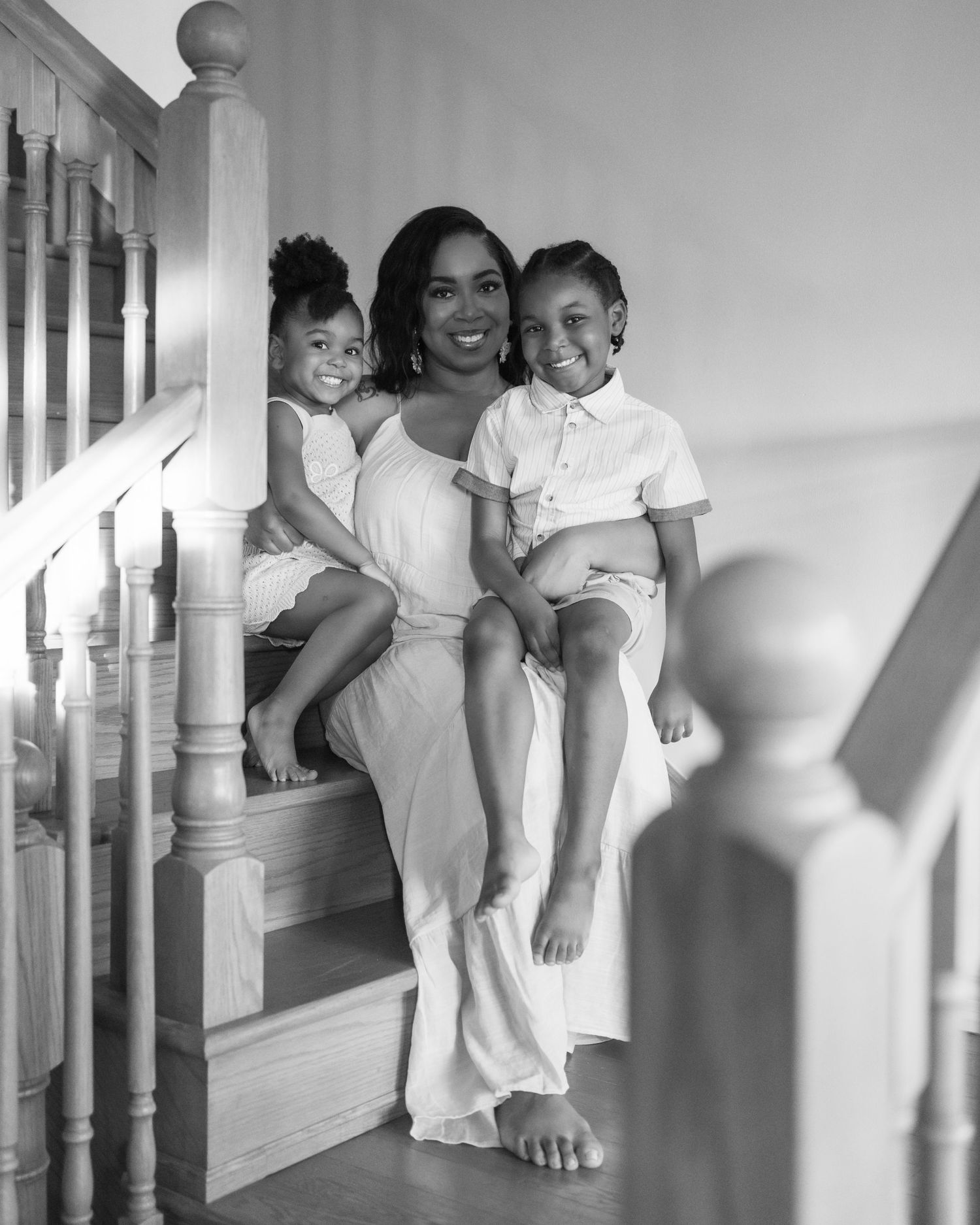 Black and white photo of a smiling woman sitting on stairs with two young children, all dressed in white.