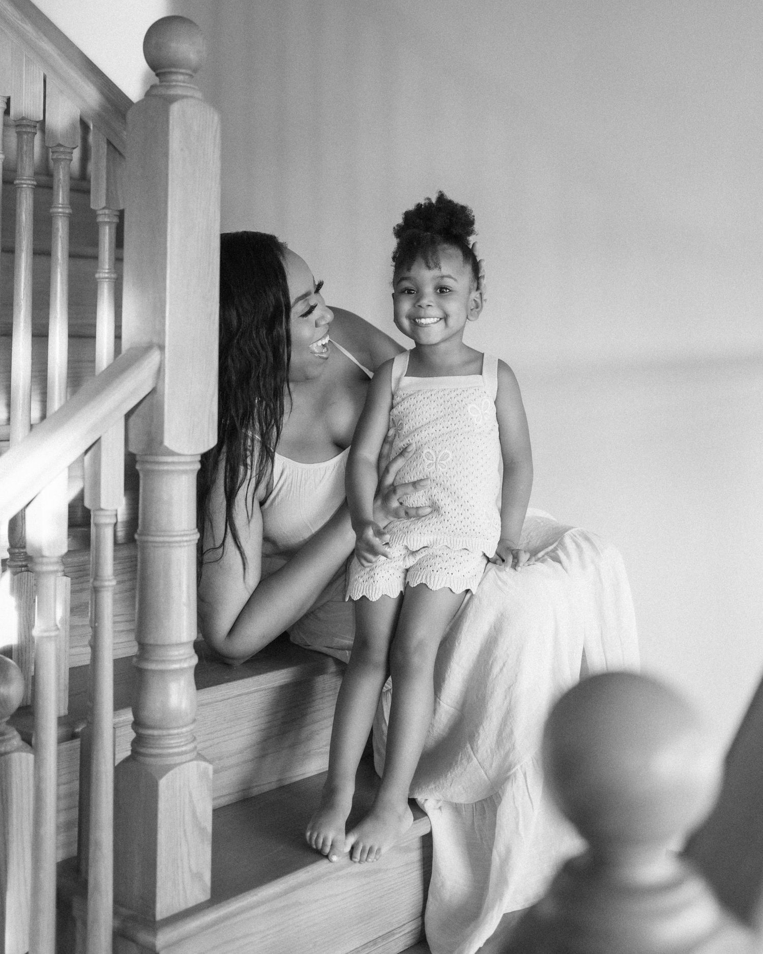 Black and white photo of a woman and young girl smiling together on wooden staircase at home.