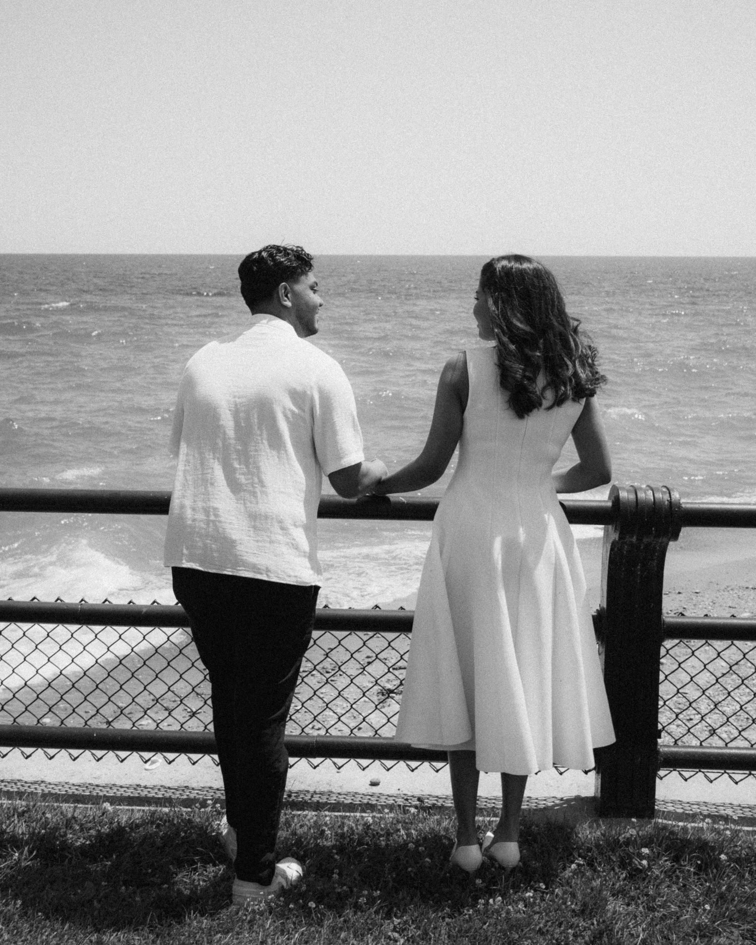 Black and white photo of a couple in white outfits holding hands while gazing at the ocean from a seaside railing.
