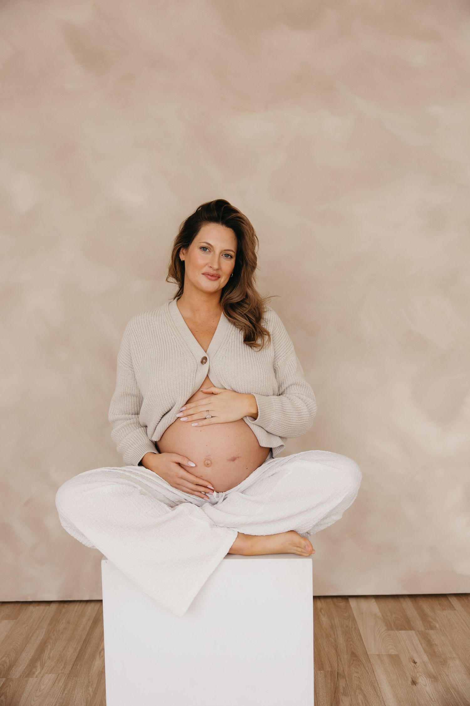 Pregnant woman in beige cardigan sitting cross-legged on white cube cradling bare bump.