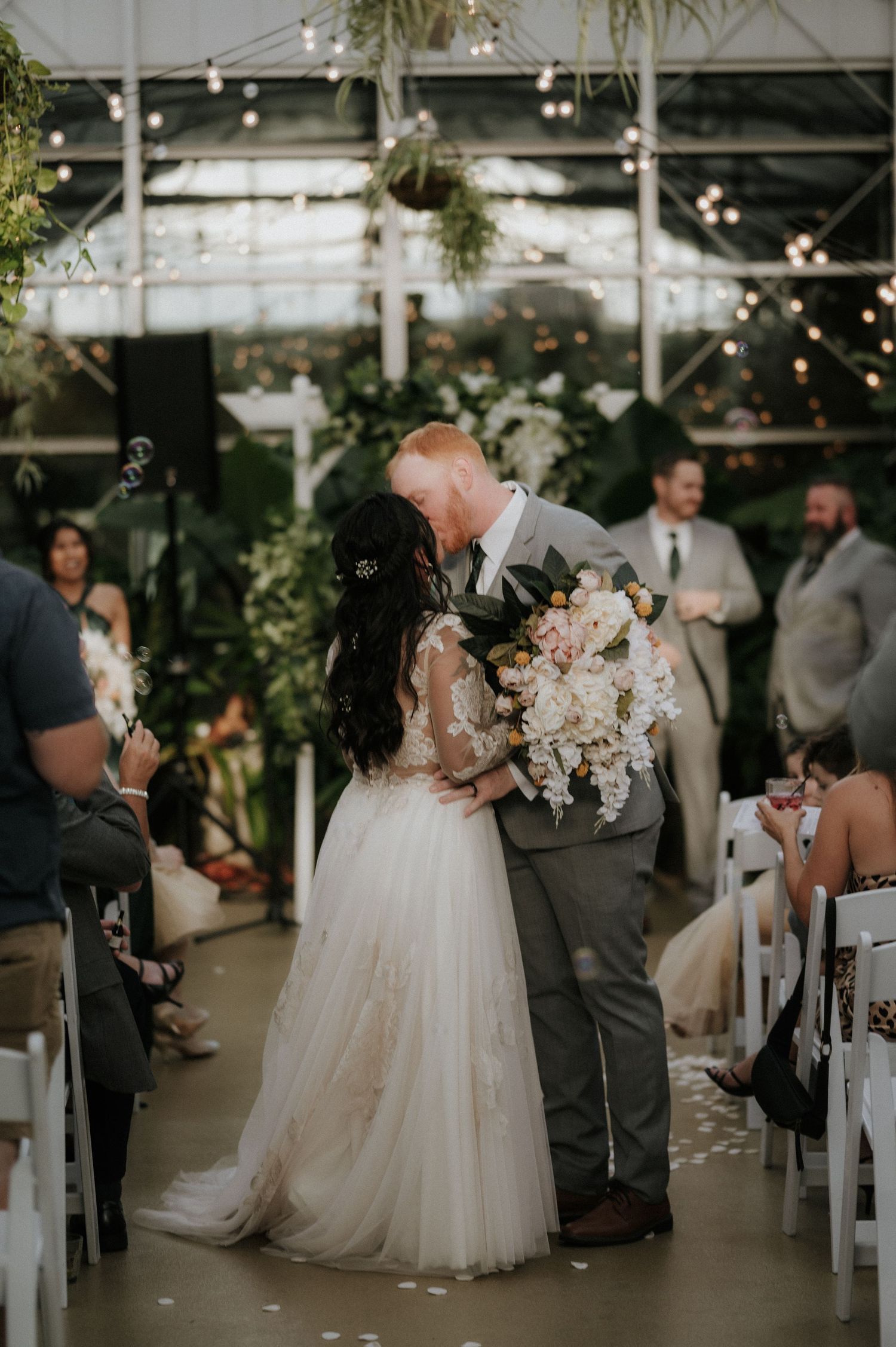 Couple sharing their first kiss during an indoor greenhouse wedding ceremony with string lights overhead.