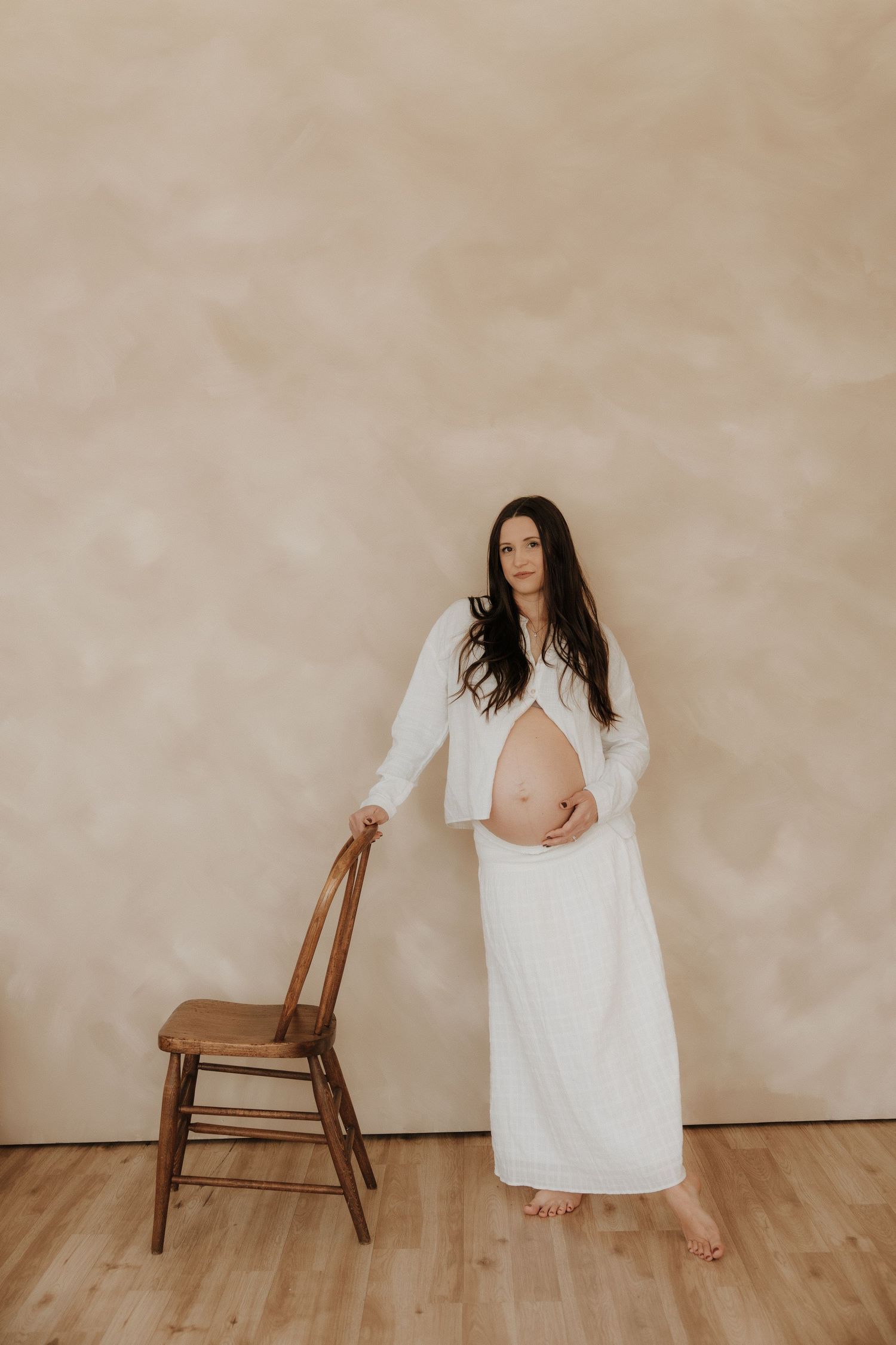 Maternity photography session featuring a white dress and wooden chair against a neutral backdrop.