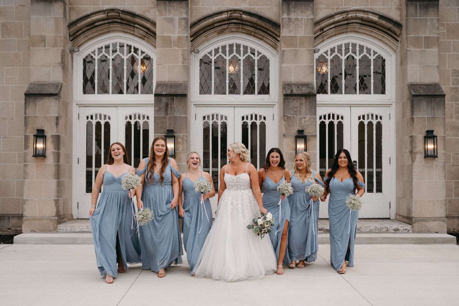 Bridal party in dusty blue gowns poses against stone church facade with arched windows.