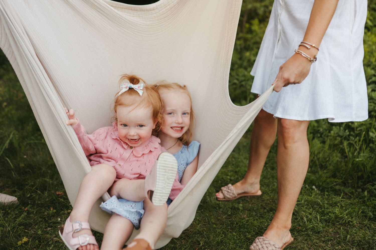 Two children laughing together while sitting in a cream-colored hammock outdoors.