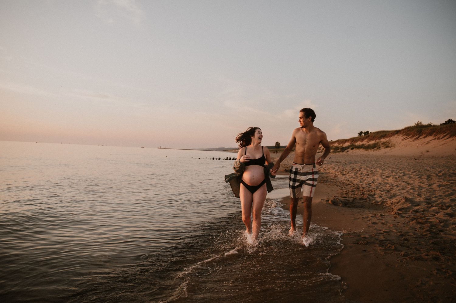 Couple enjoying a playful moment walking through shallow water on a beach at sunset.