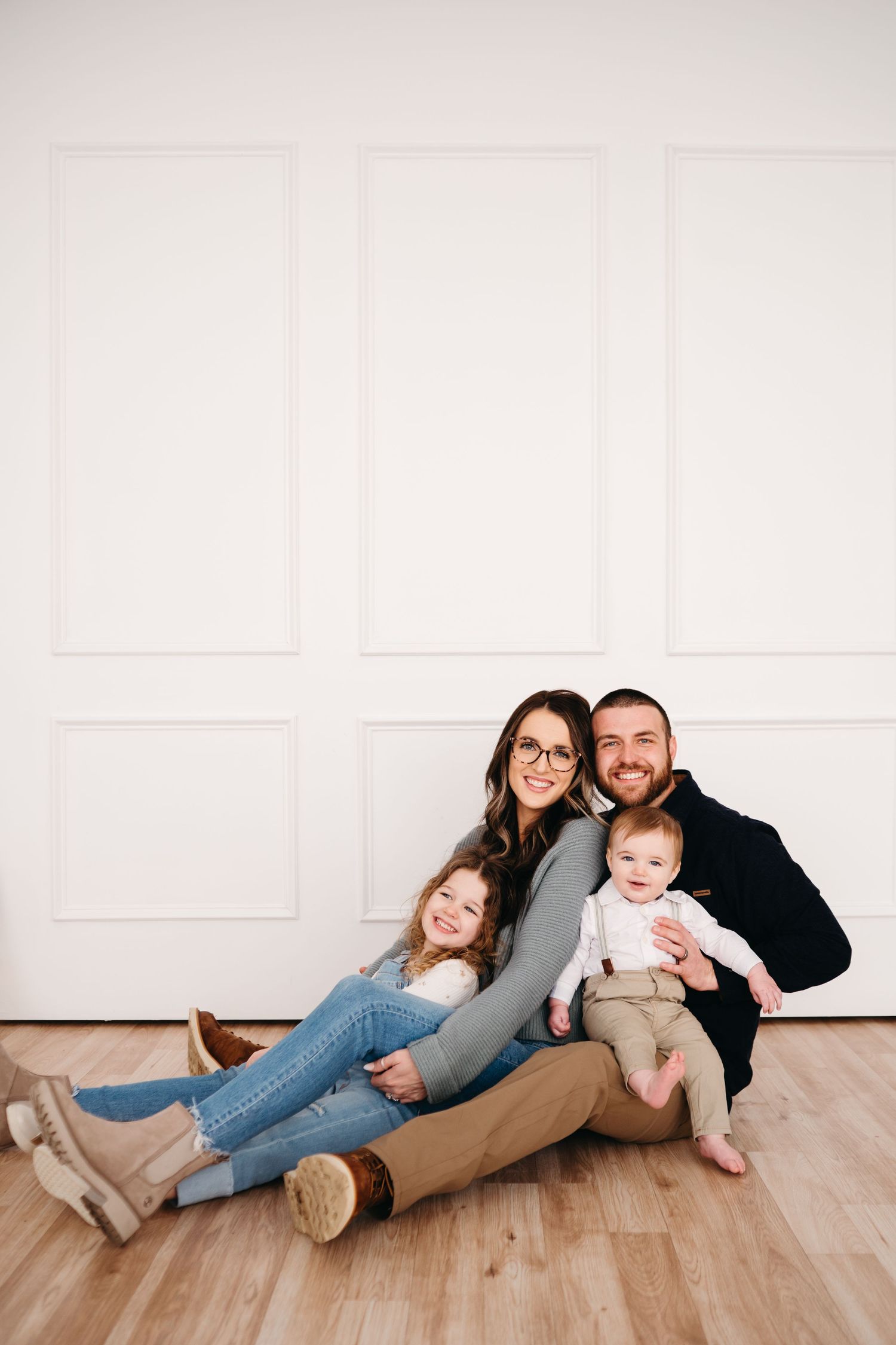 Candid family portrait sitting on wooden floor against white paneled wall.