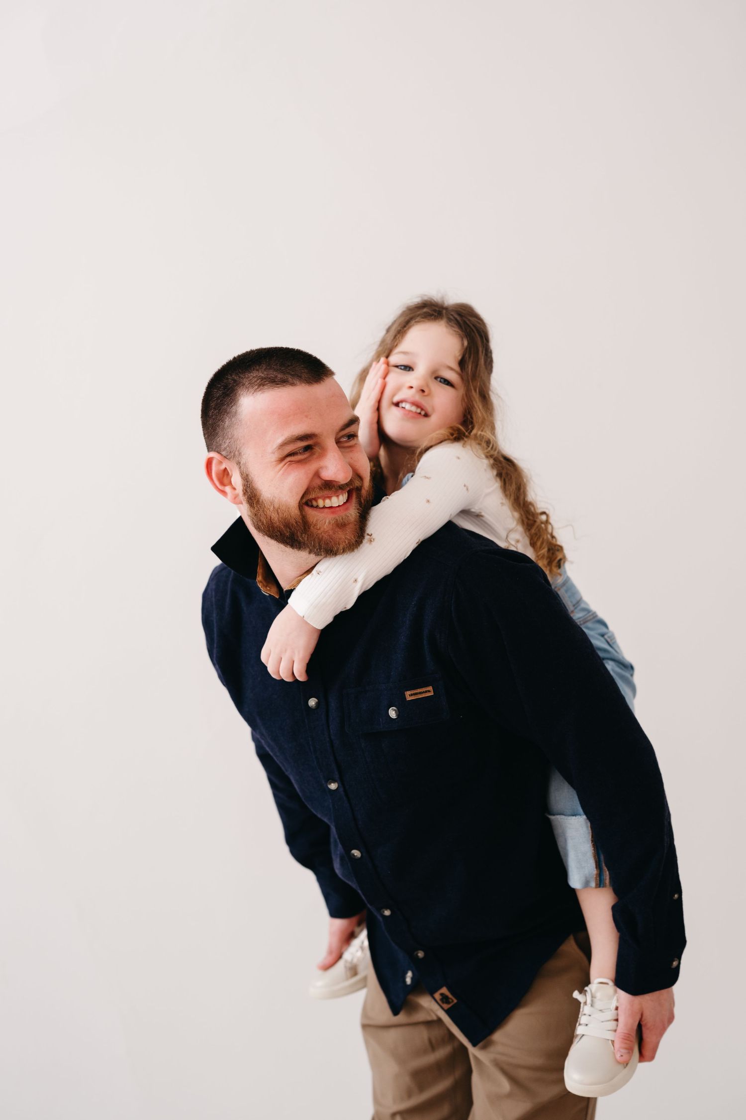 Playful piggyback ride against a white background as child in white sweater hugs around neck with joyful expressions.