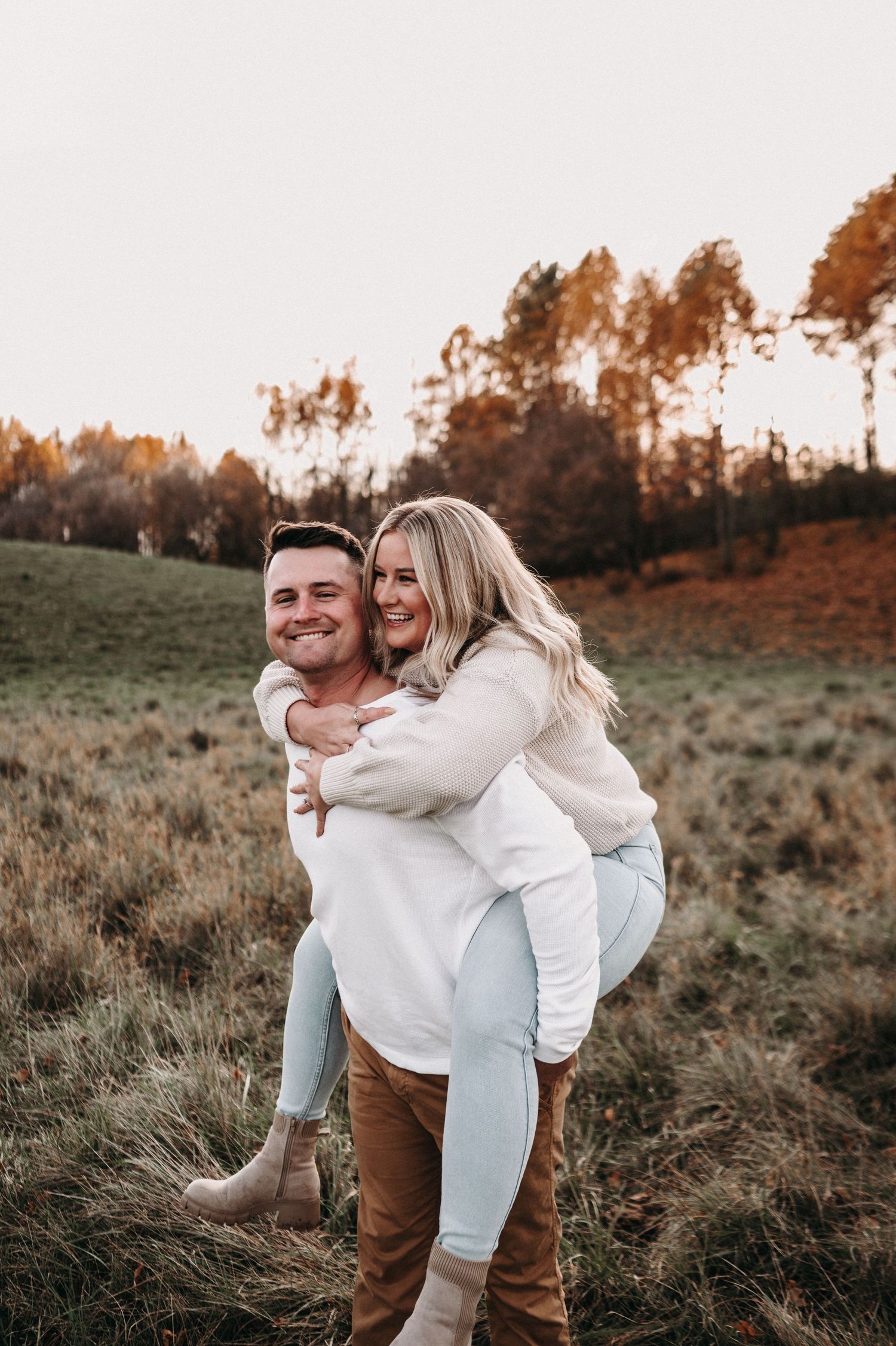 Happy couple enjoying piggyback ride outdoors against autumn trees at sunset.