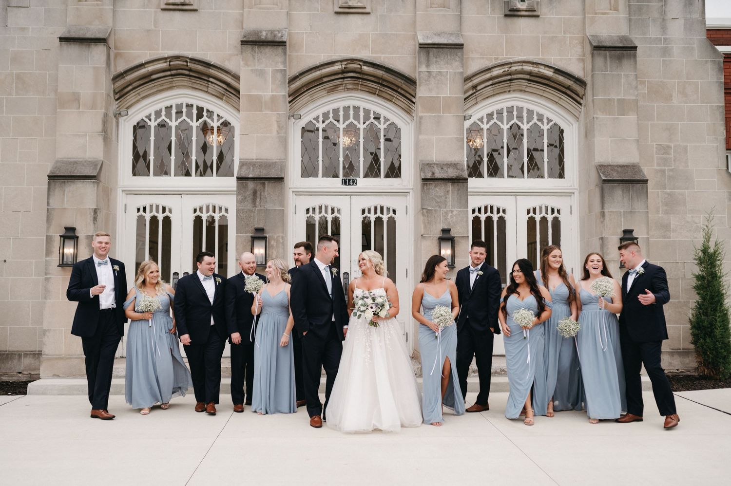 Wedding party in blue dresses and black suits poses casually against stone church background.