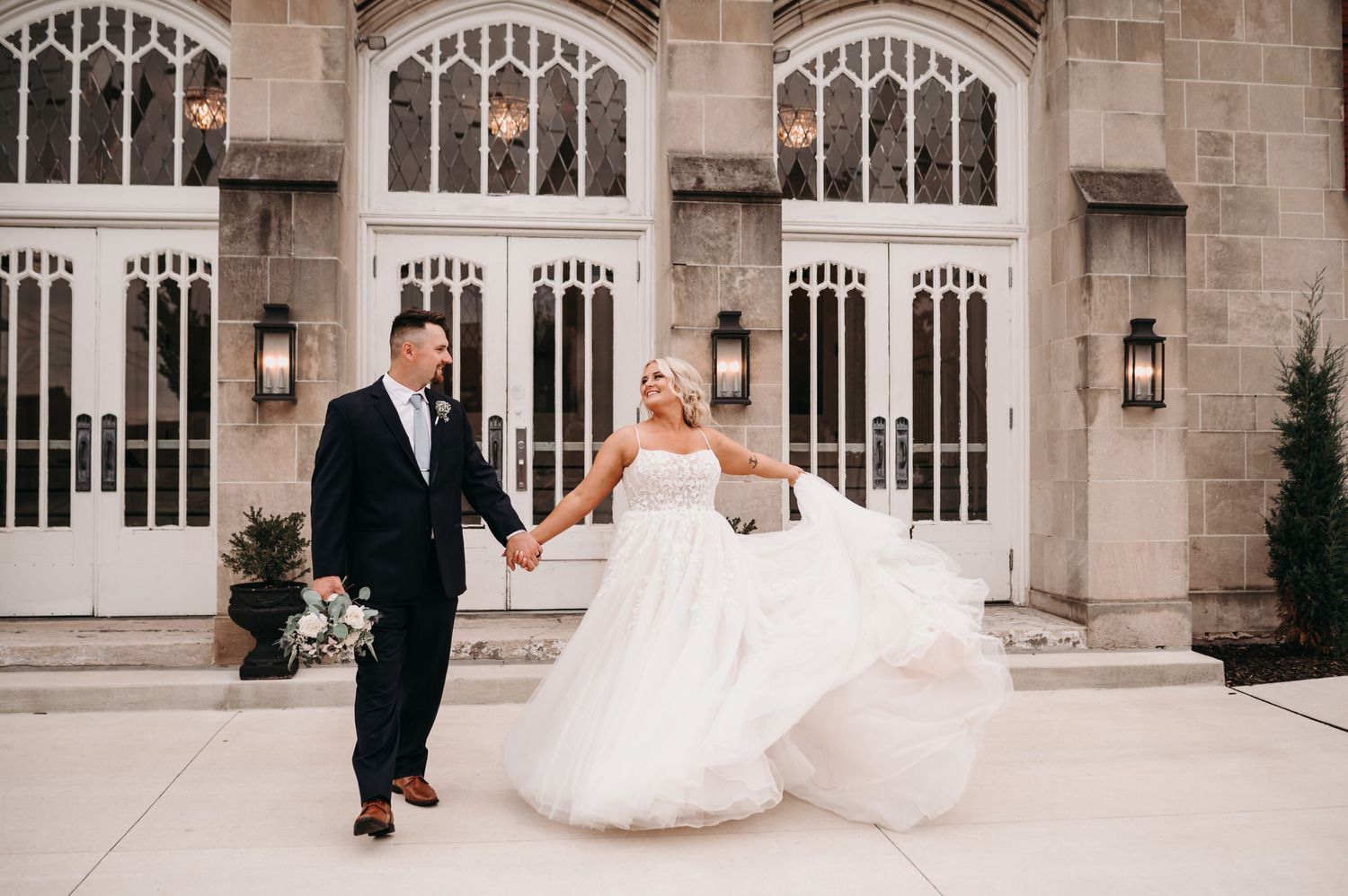 A couple shares a joyful moment outside a Gothic-style venue with their wedding attire flowing in the wind.