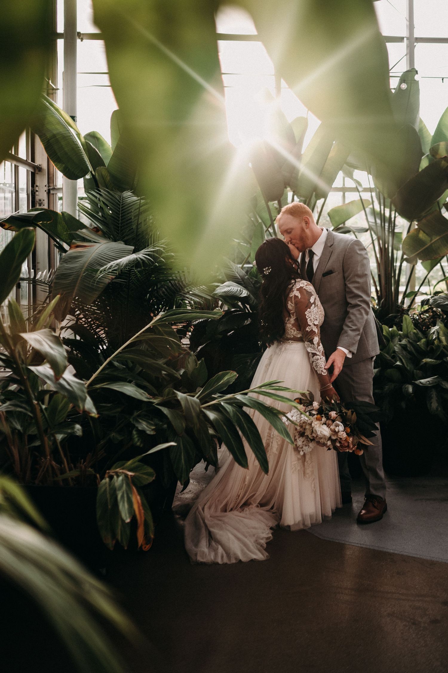 Intimate wedding portrait through tropical leaves in a sunlit greenhouse setting.