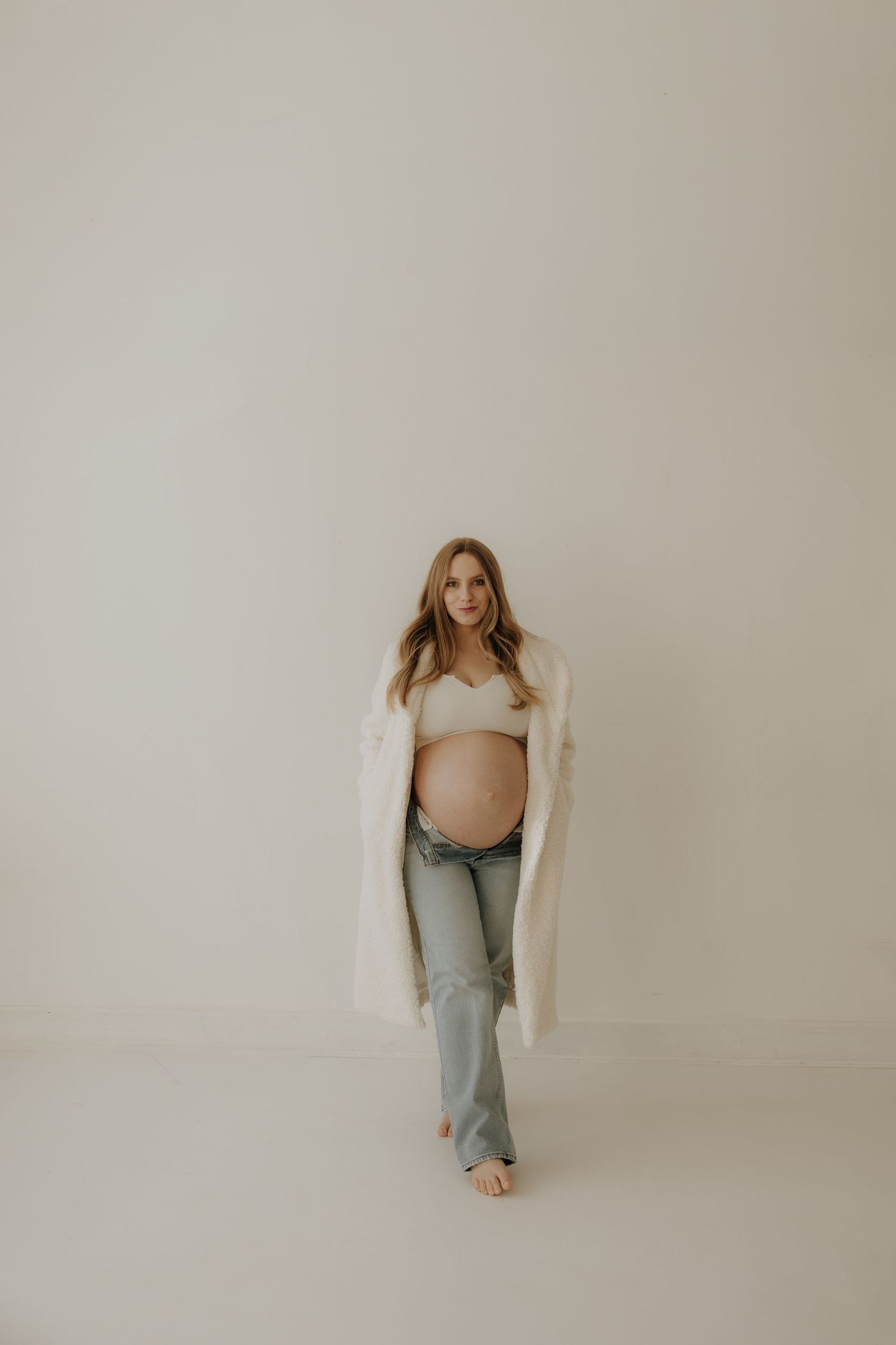 Expectant mother in light cardigan and jeans walking against white backdrop in studio setting.