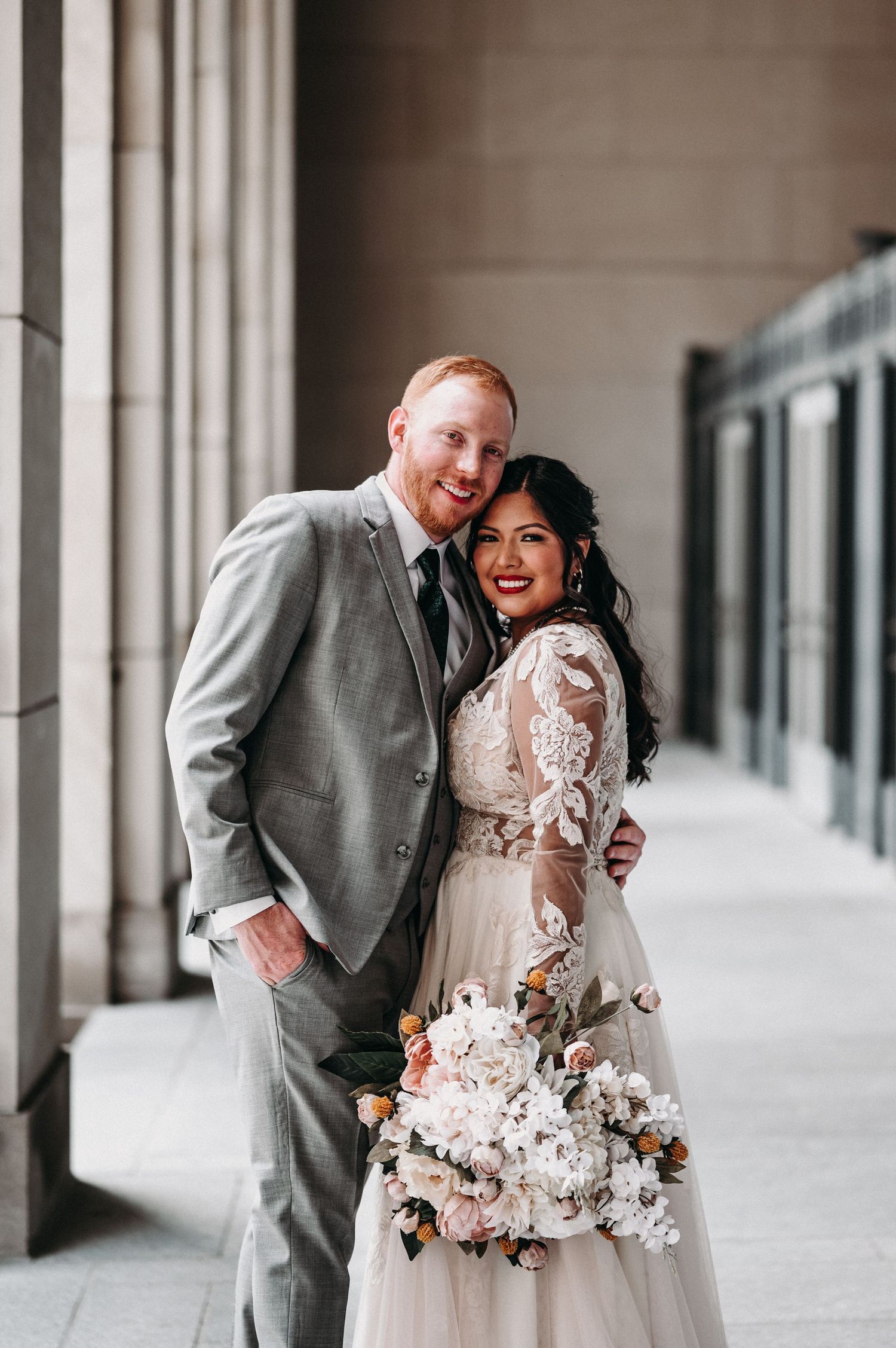 A classic wedding portrait featuring a couple in gray suit and lace dress holding a pink floral bouquet.