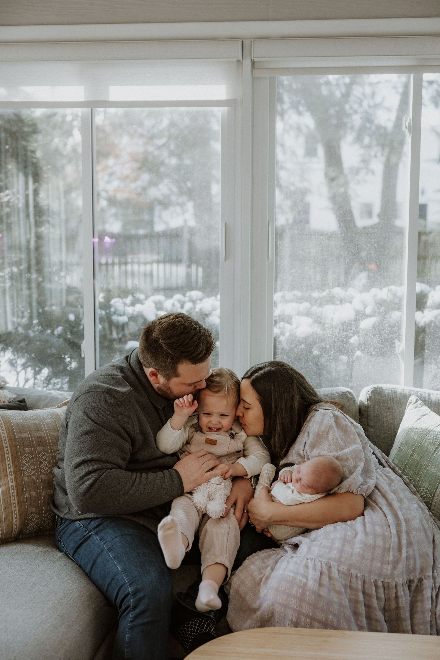 A tender family moment on a gray couch with snowy winter scene visible through windows.