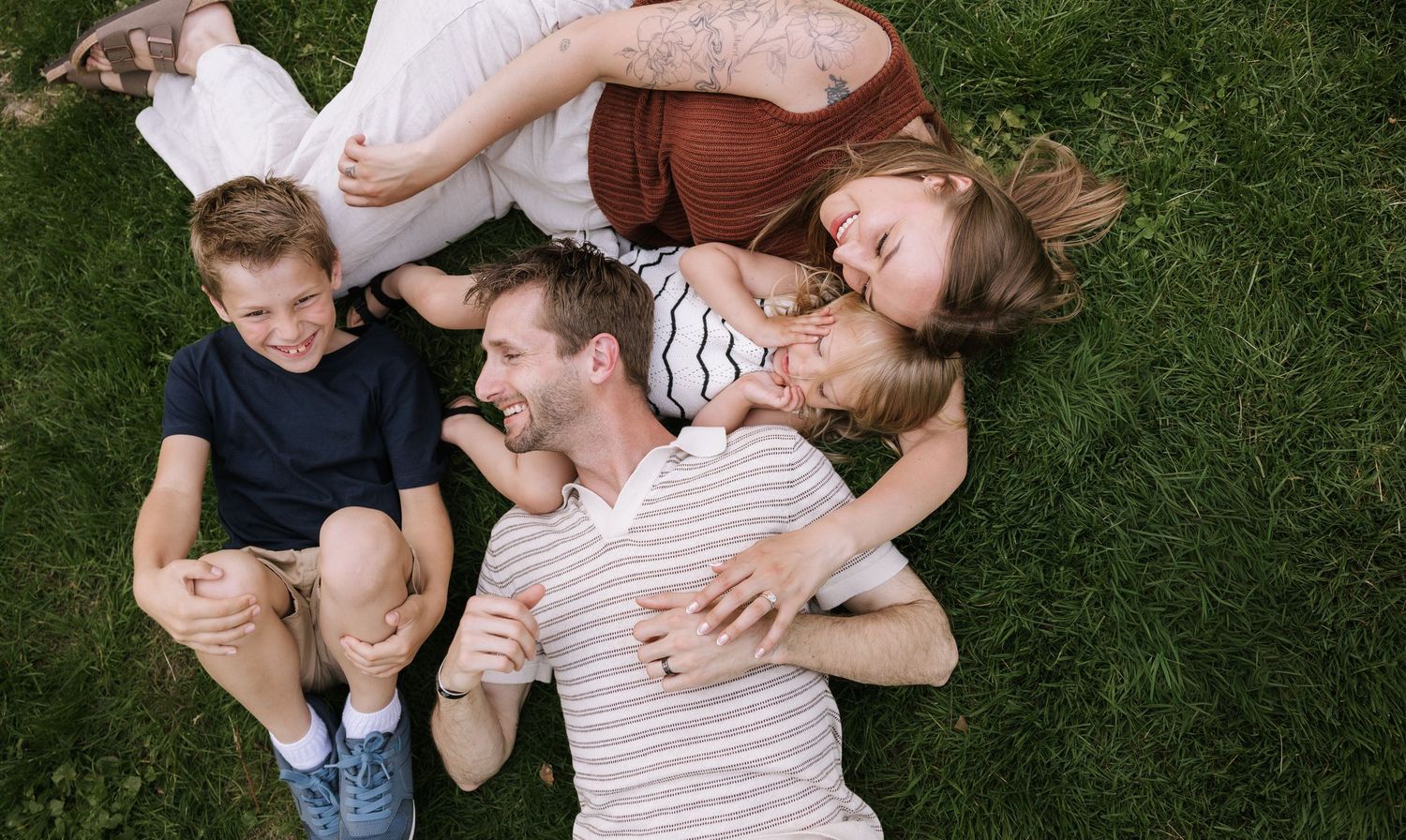 Happy family of four lying on green grass together, laughing and enjoying a sunny day outdoors.