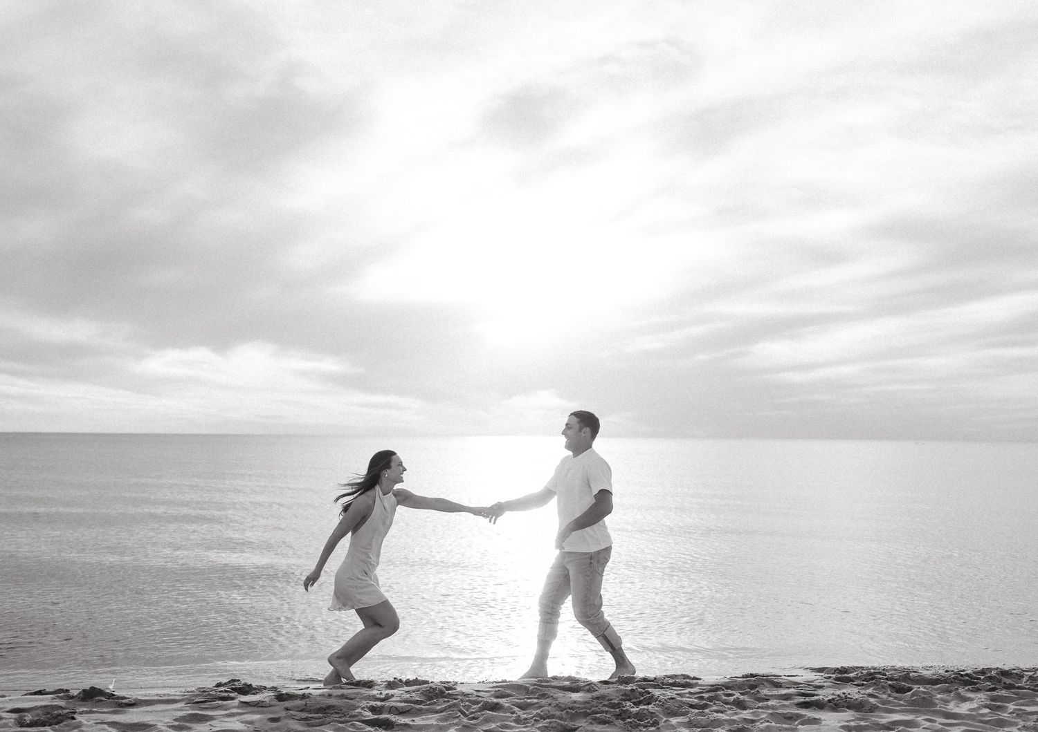 Black and white photo of a couple joyfully holding hands on a sunlit beach.
