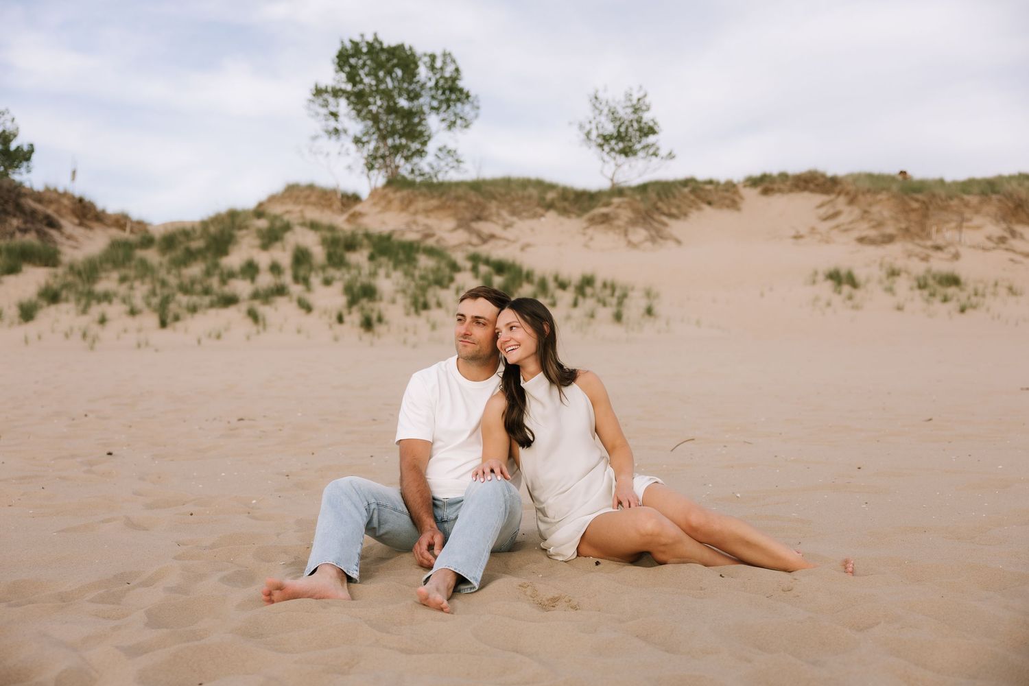 Happy couple sitting together on sandy dunes with trees in the background during an outdoor engagement session.