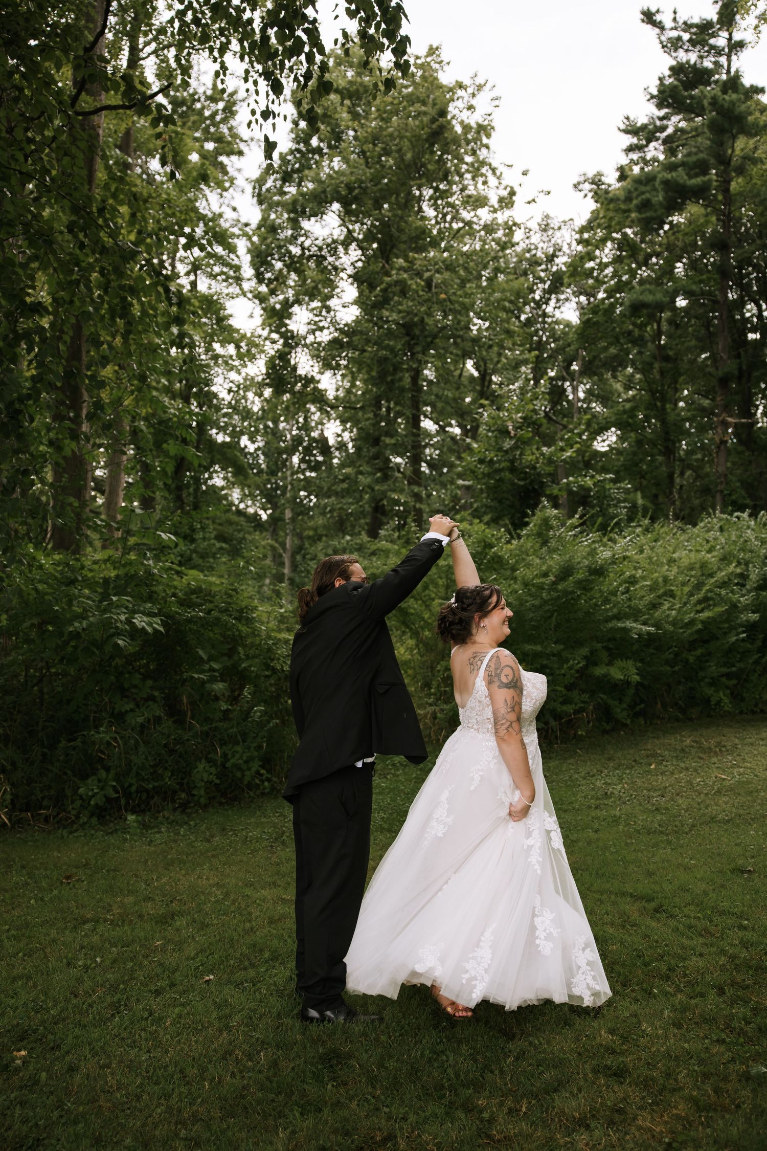 Dancing couple in wedding attire twirling on green grass with trees in background.